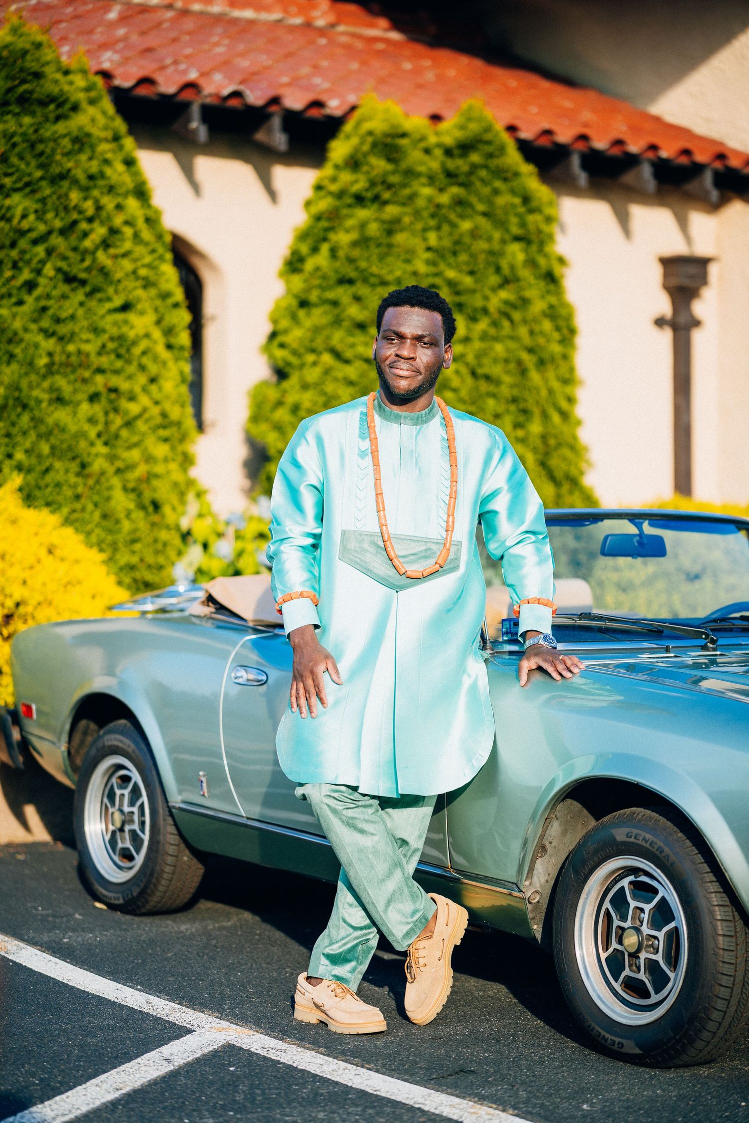 Groom in teal African attire leans on vintage convertible car.