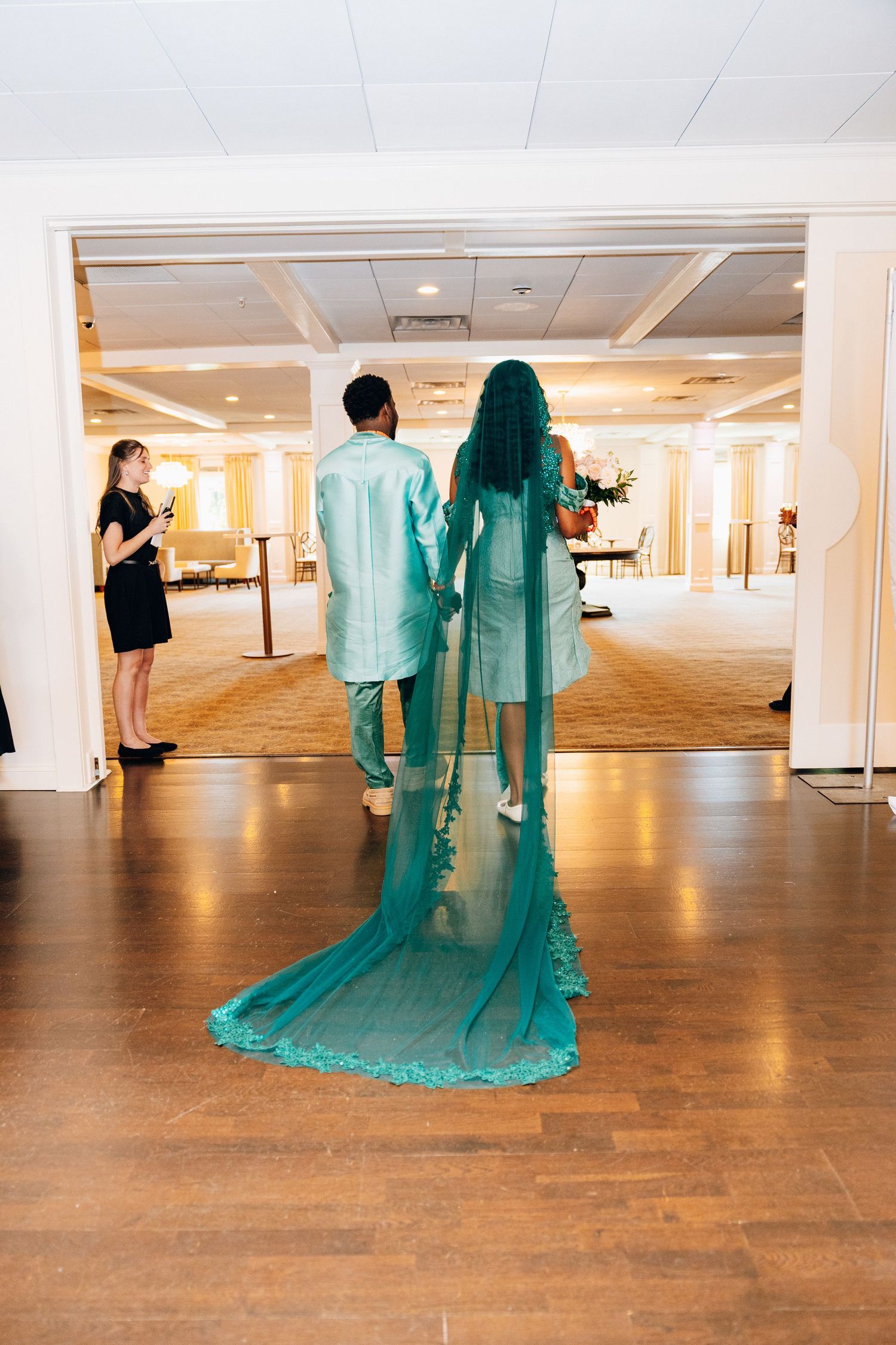 Bride and groom in matching teal outfits walk hand in hand into wedding reception hall.