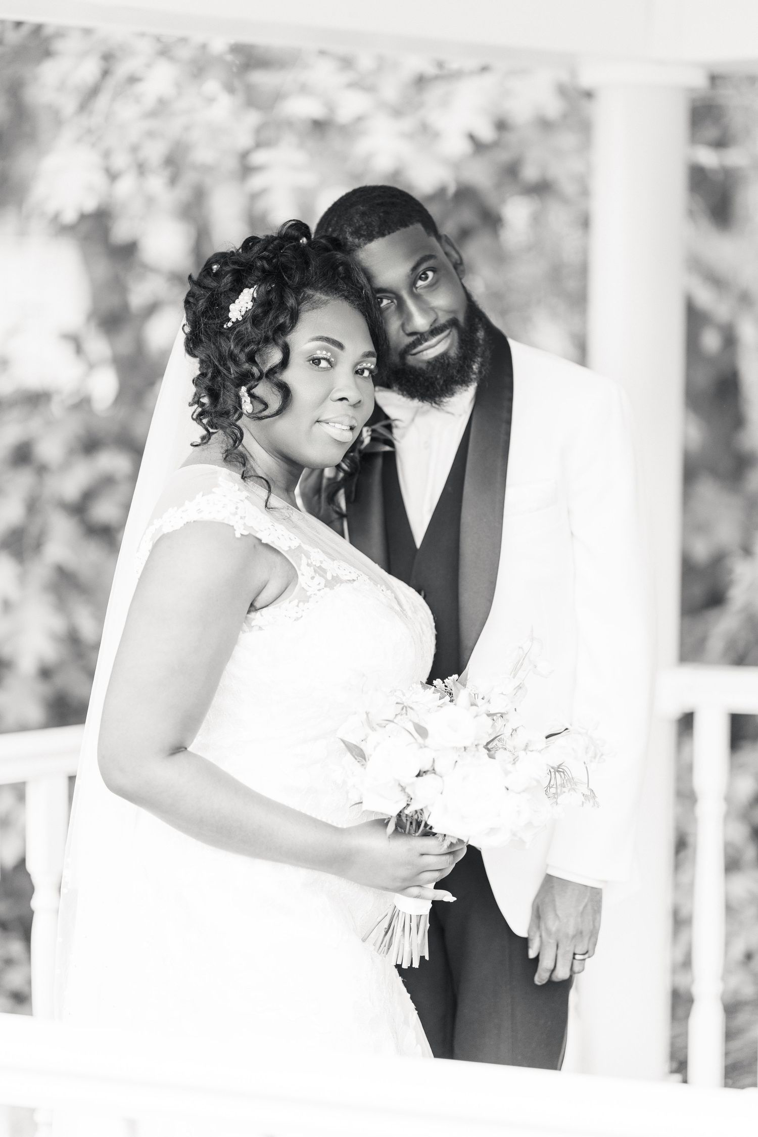 Black and white wedding portrait of a smiling bride in lace gown with bouquet and groom in white tuxedo on a porch.