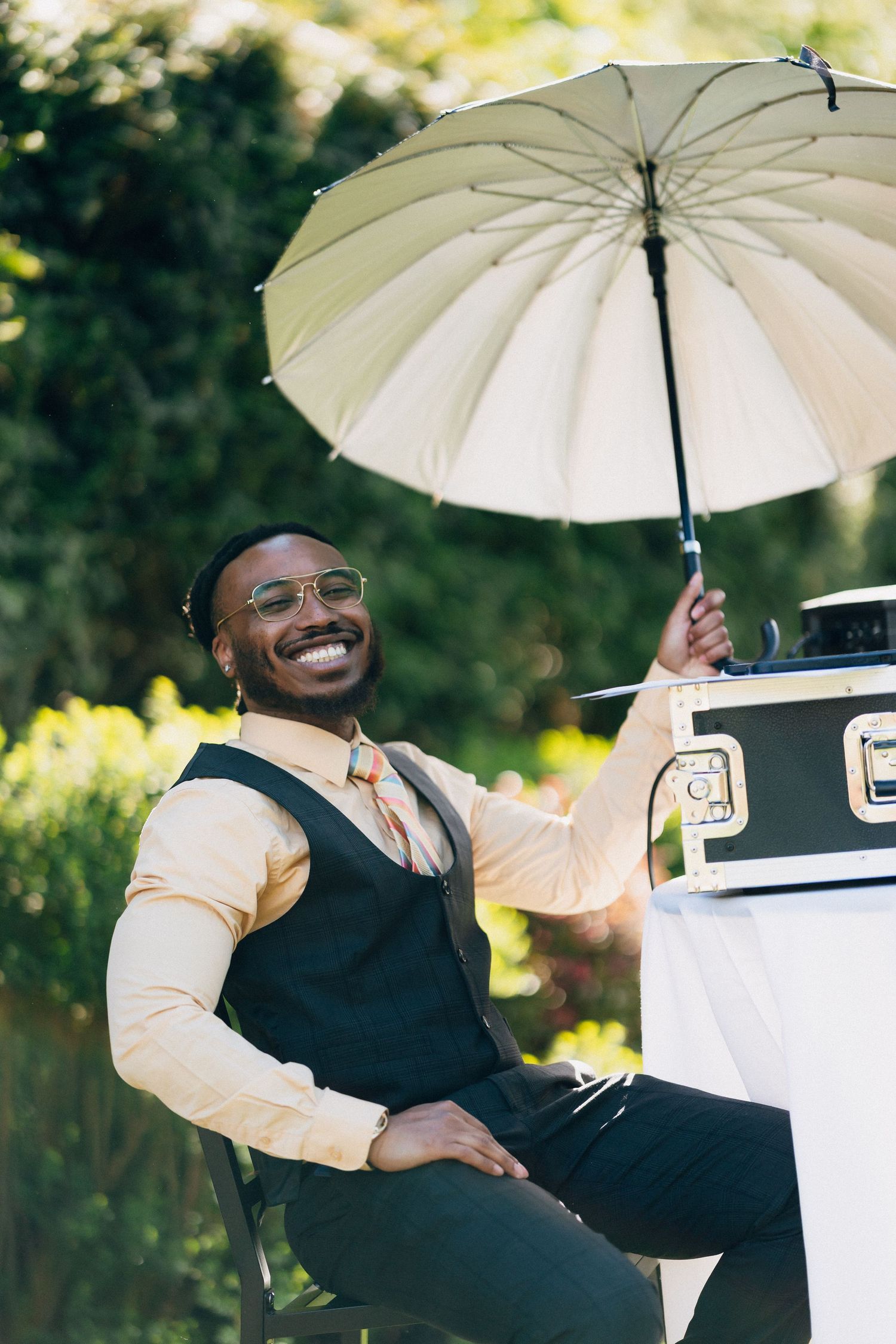 Smiling Black man in vest and glasses holds white umbrella outdoors at garden event.