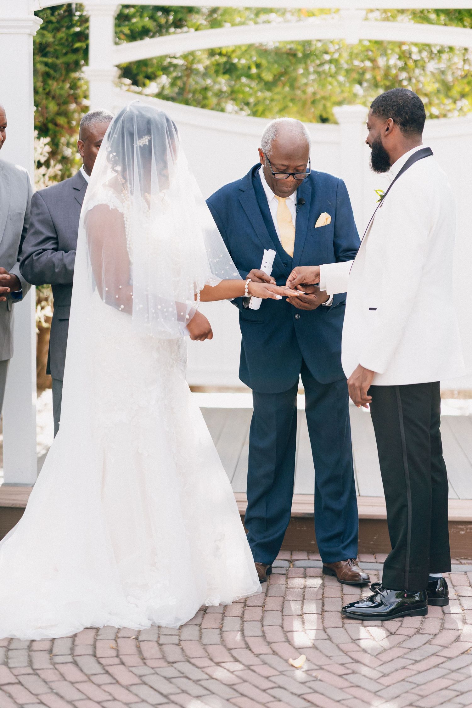 Bride in white gown and veil exchanging rings with groom in white tuxedo at outdoor ceremony.