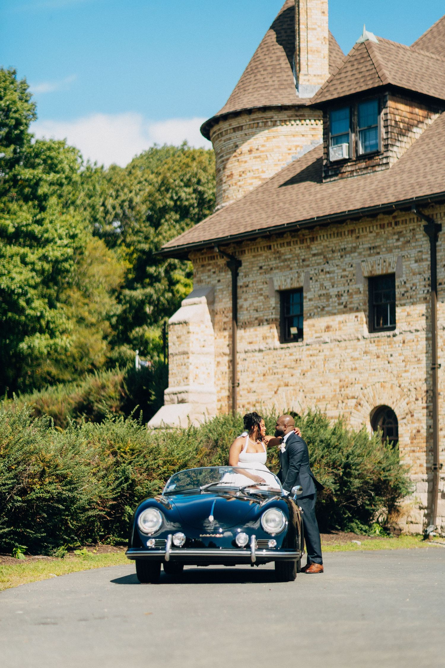 Wedding couple posing with a classic navy Porsche convertible in front of a stone castle venue.