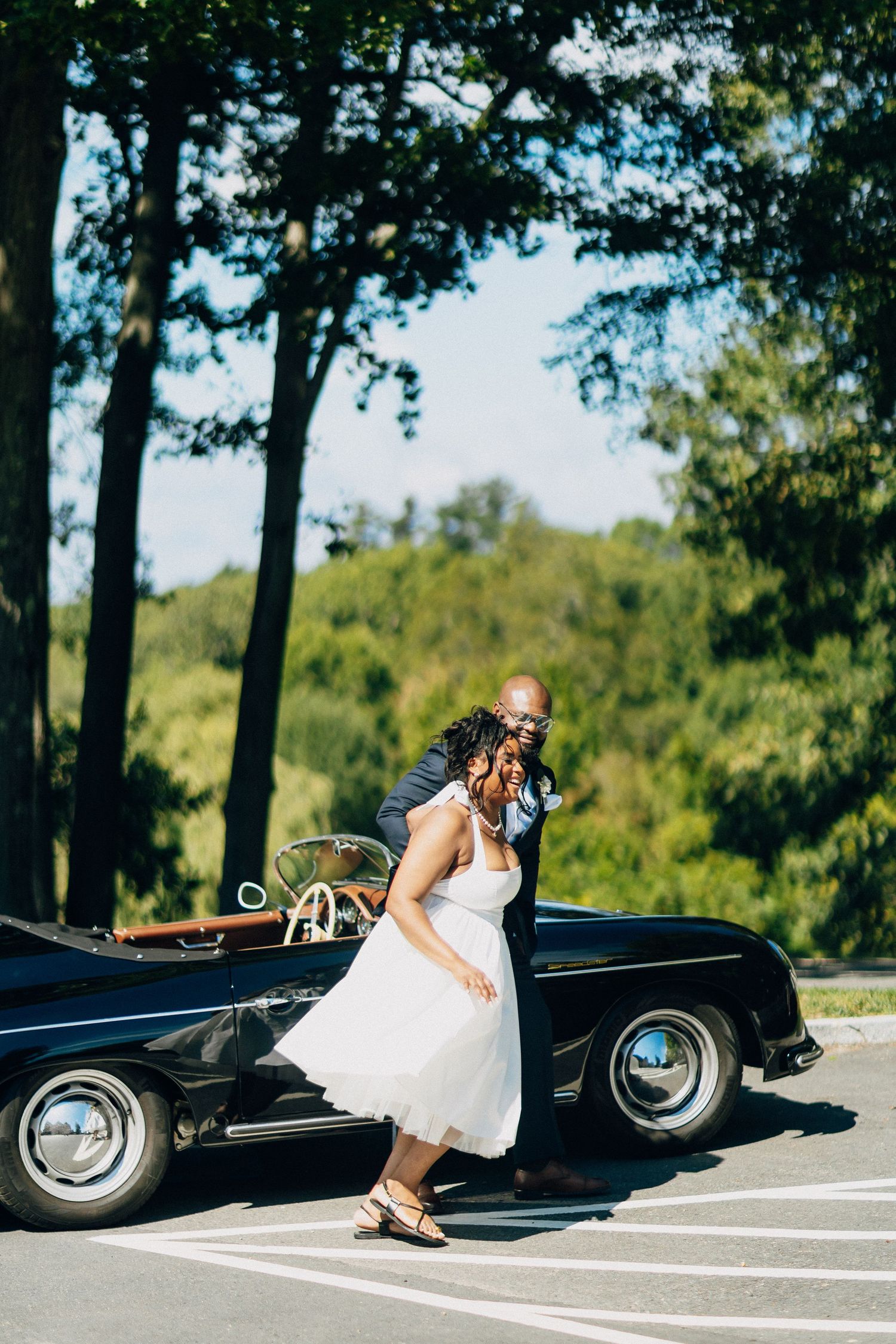 A groom playfully dips his bride beside a classic black convertible under sunny skies.