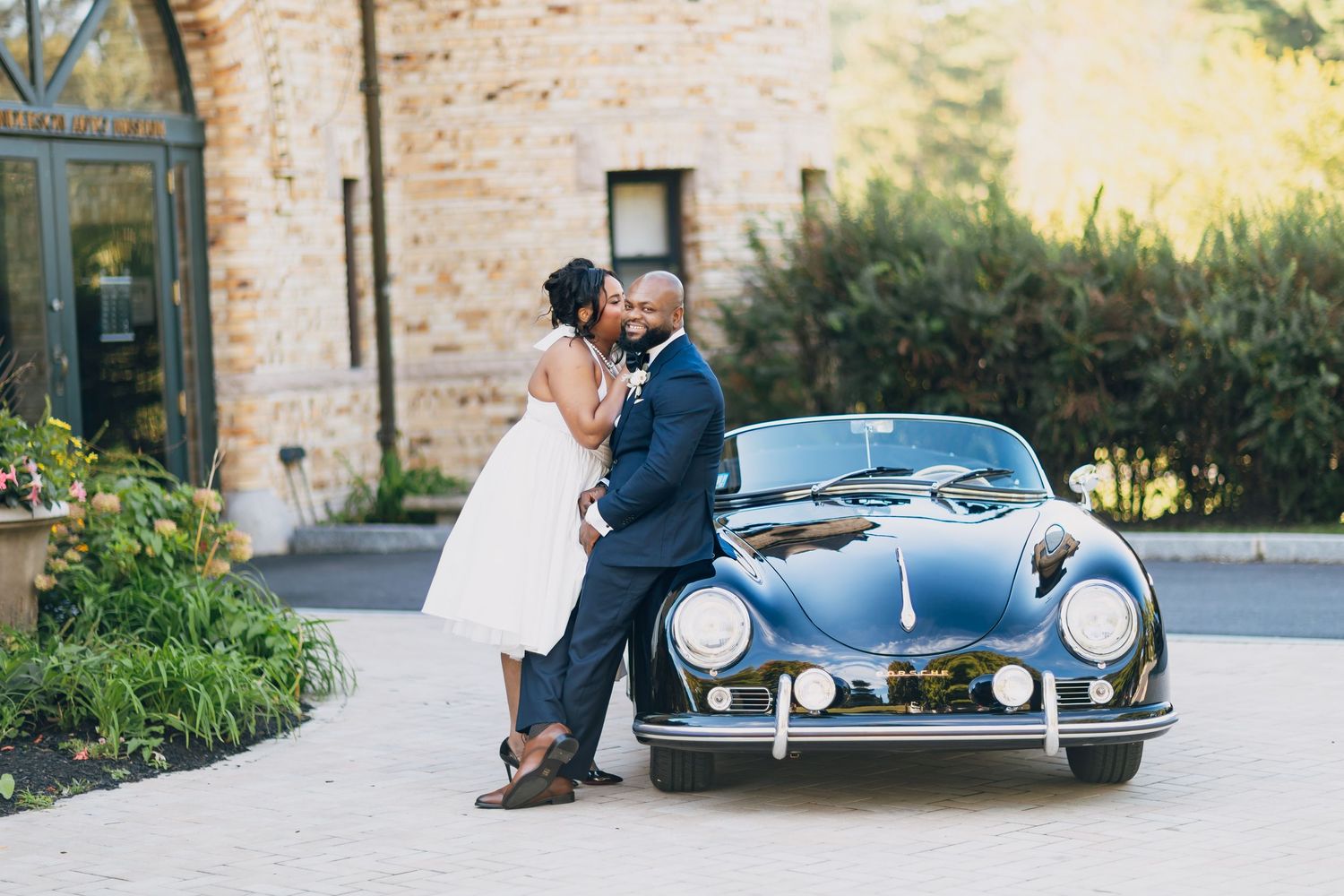 A couple shares a romantic kiss beside a classic navy Porsche at their elegant outdoor wedding venue.