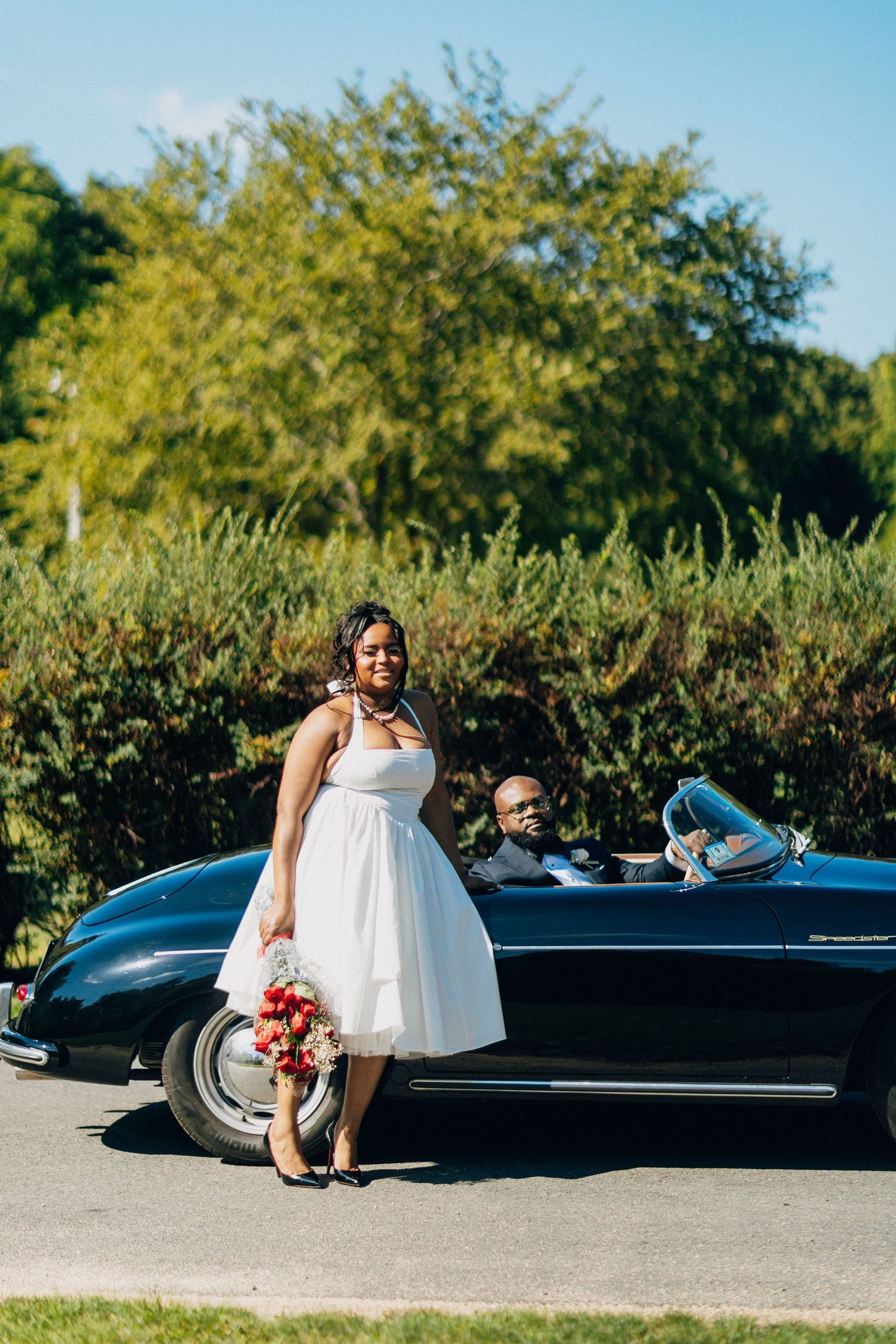 Smiling bride holds red bouquet beside a classic black convertible as groom sits in the drivers seat.