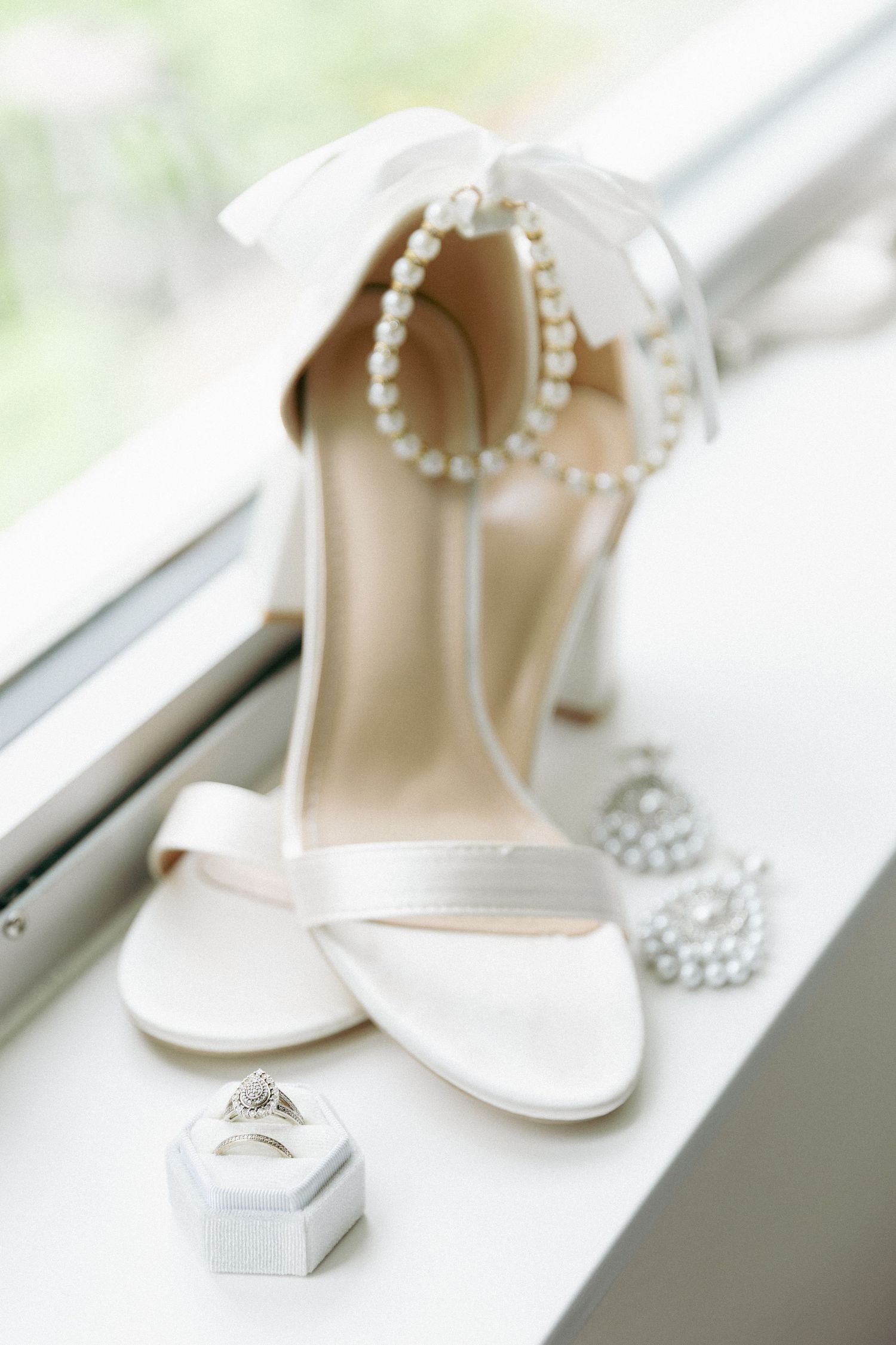 White bridal heels with pearl ankle strap and bow displayed with diamond engagement ring on white surface.