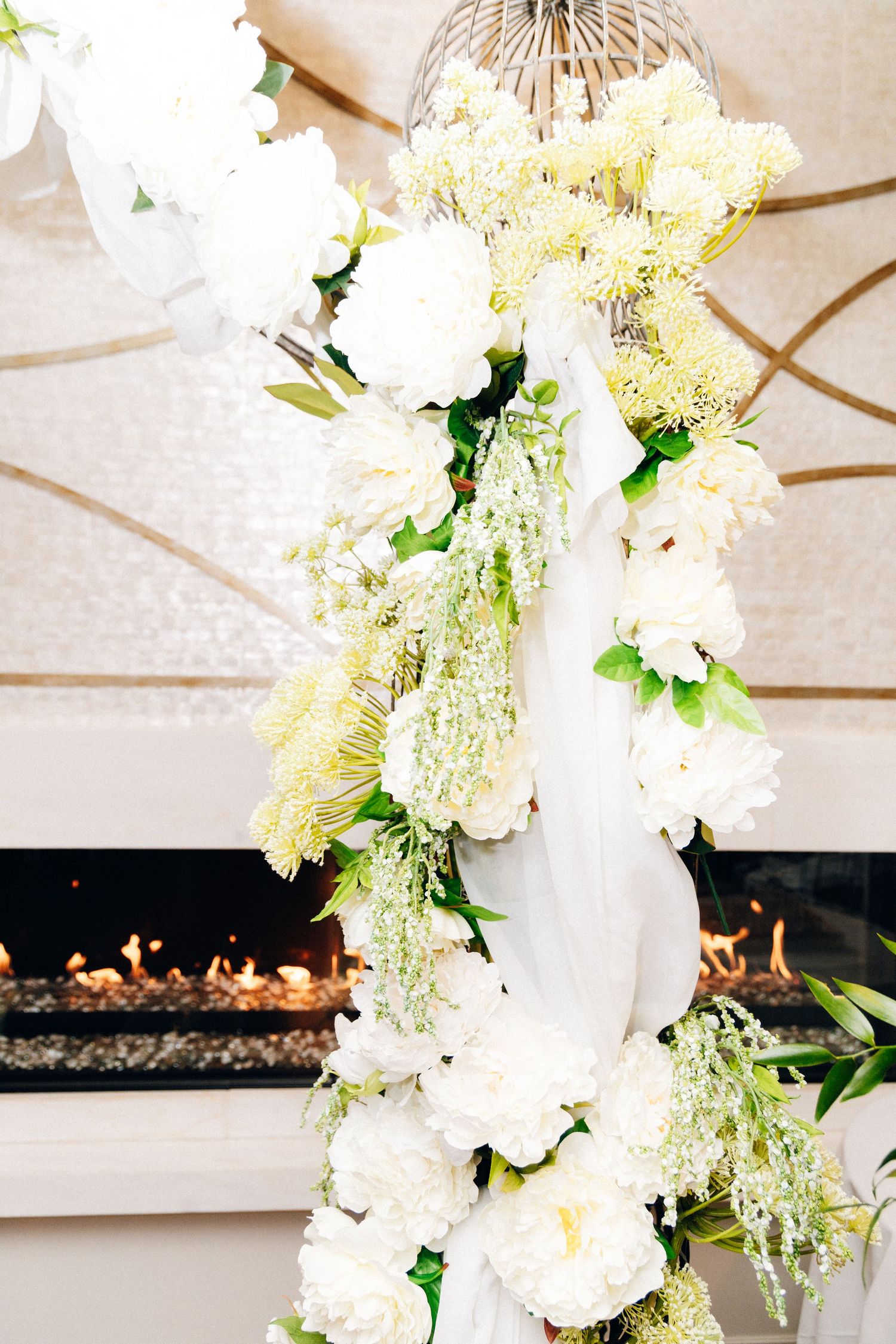Elegant white floral wedding arch with peonies and greenery draped over a modern fireplace.