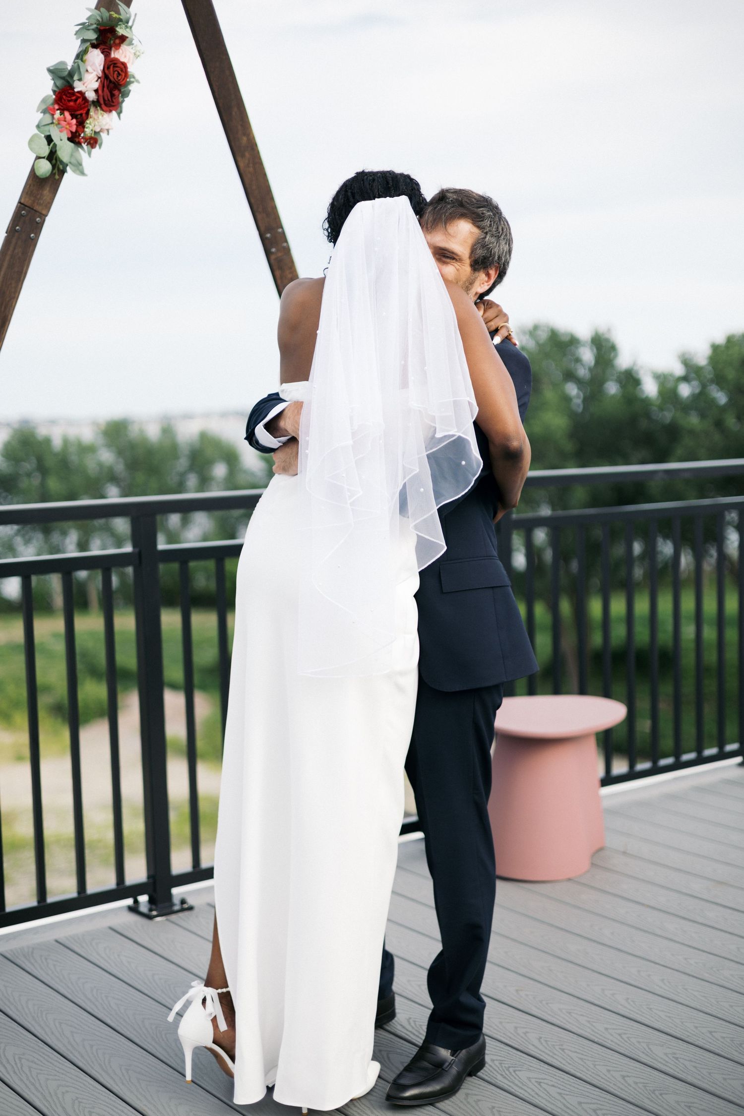 Interracial couple embracing at outdoor wedding ceremony on deck with floral arch, bride in white gown and veil.