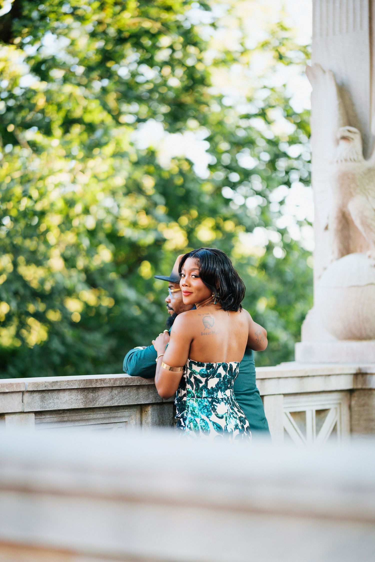 Elegant couple embracing at a stone monument in a lush green park, woman in floral green gown looking back.