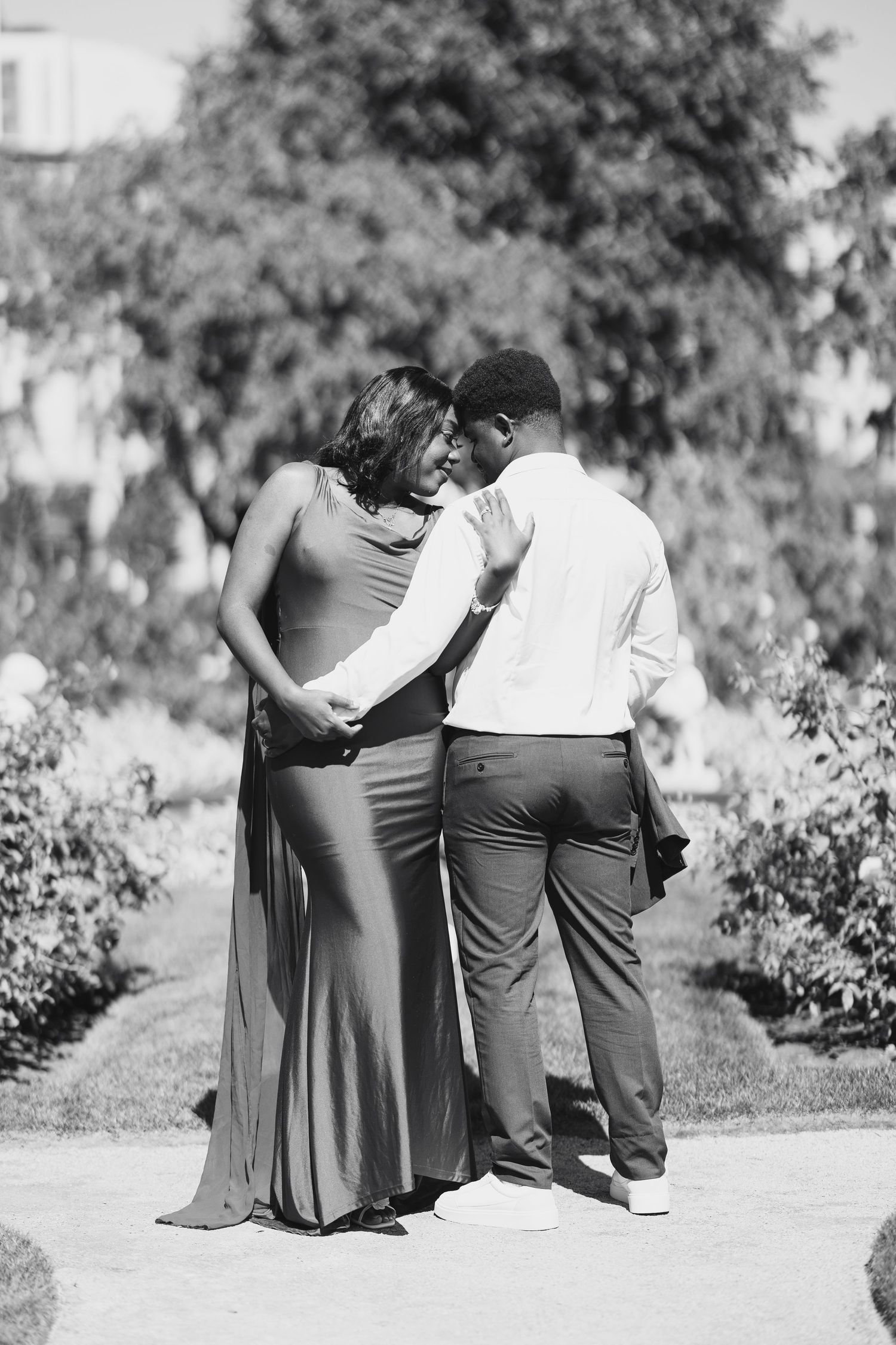 Black and white photo of a romantic couple sharing an intimate moment on a garden pathway.