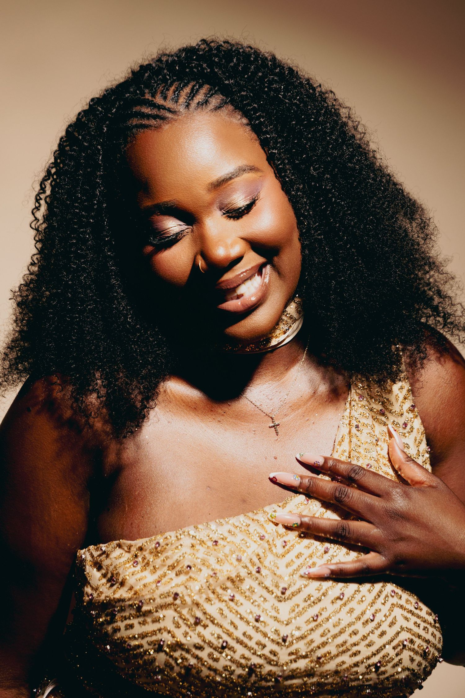 Smiling Black woman with curly braided hair wearing a gold sequin dress and cross necklace poses gracefully.