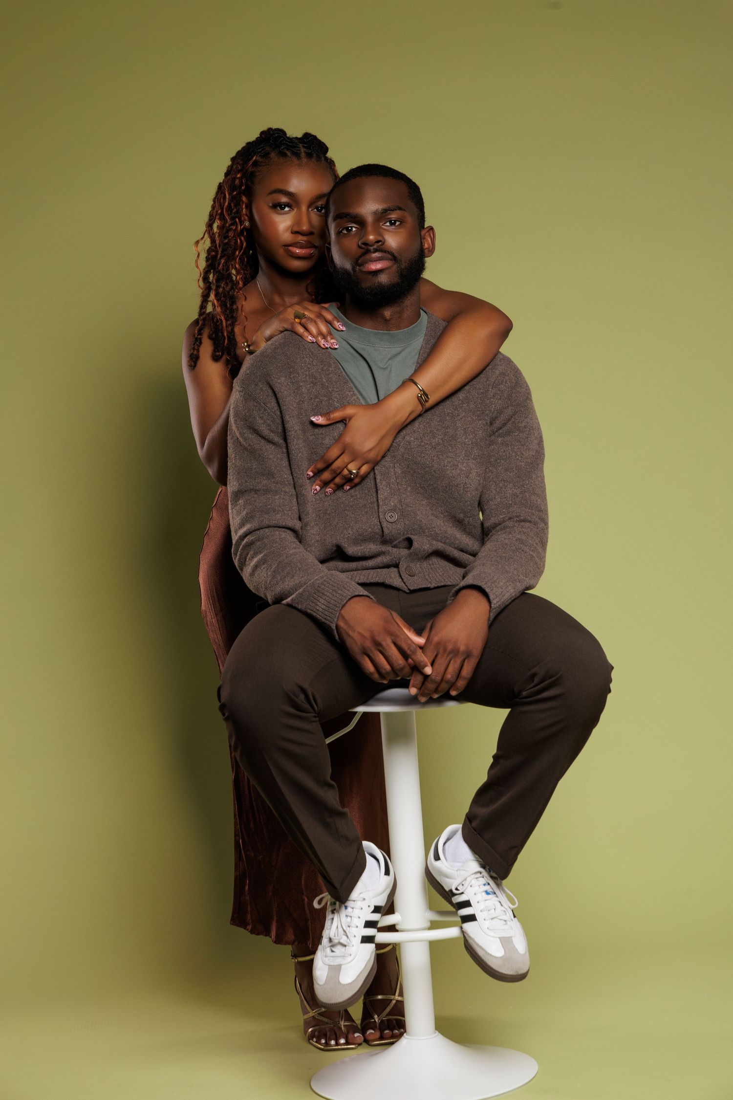 A couple poses together in a studio, woman embracing man seated on white stool, green background.