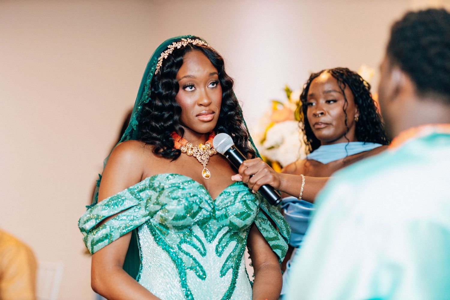 Young woman in green sequin off-shoulder gown speaking into microphone at formal event.