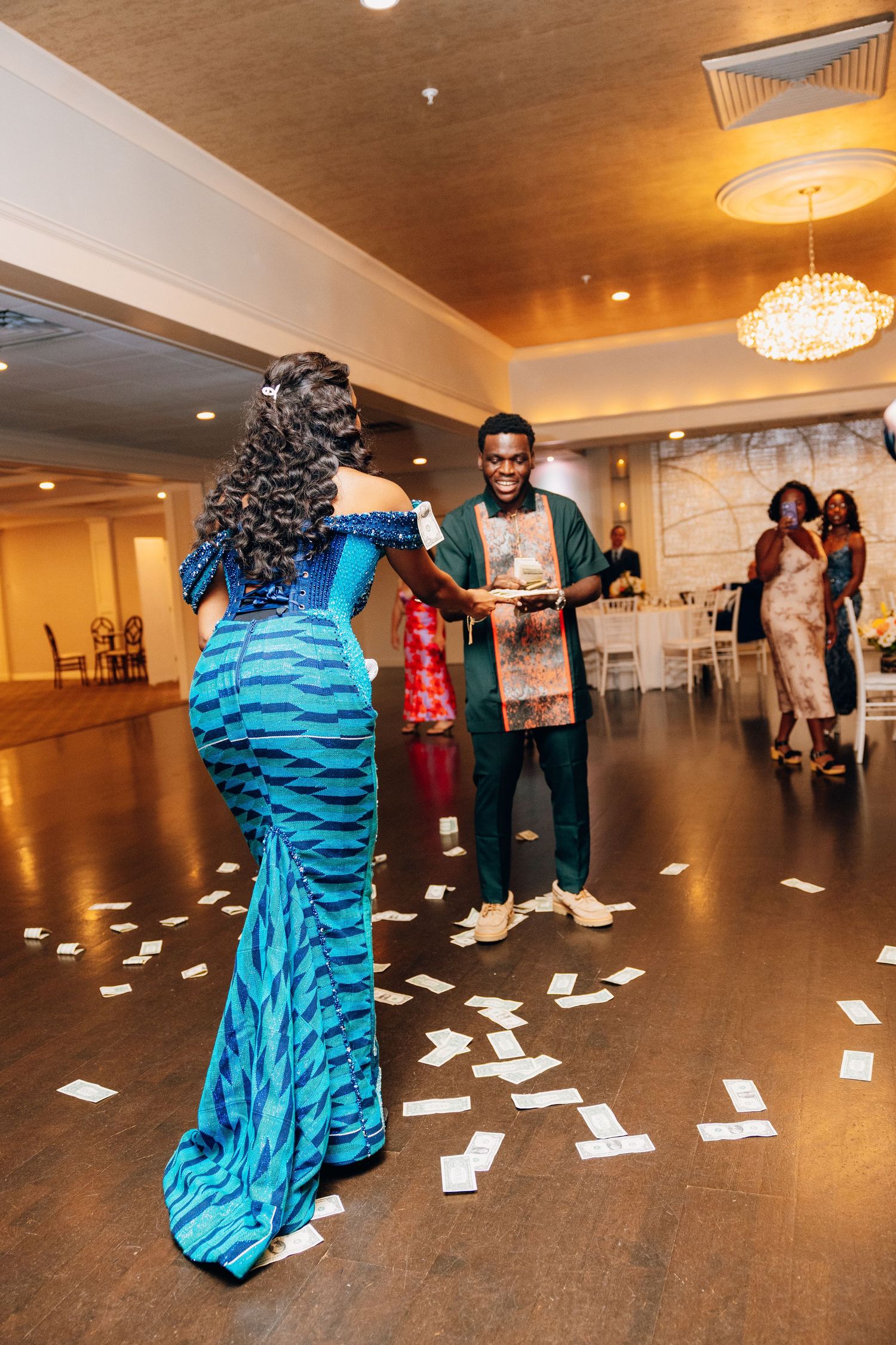 Man in green suit sprays money at woman in blue mermaid dress on dance floor at elegant reception.