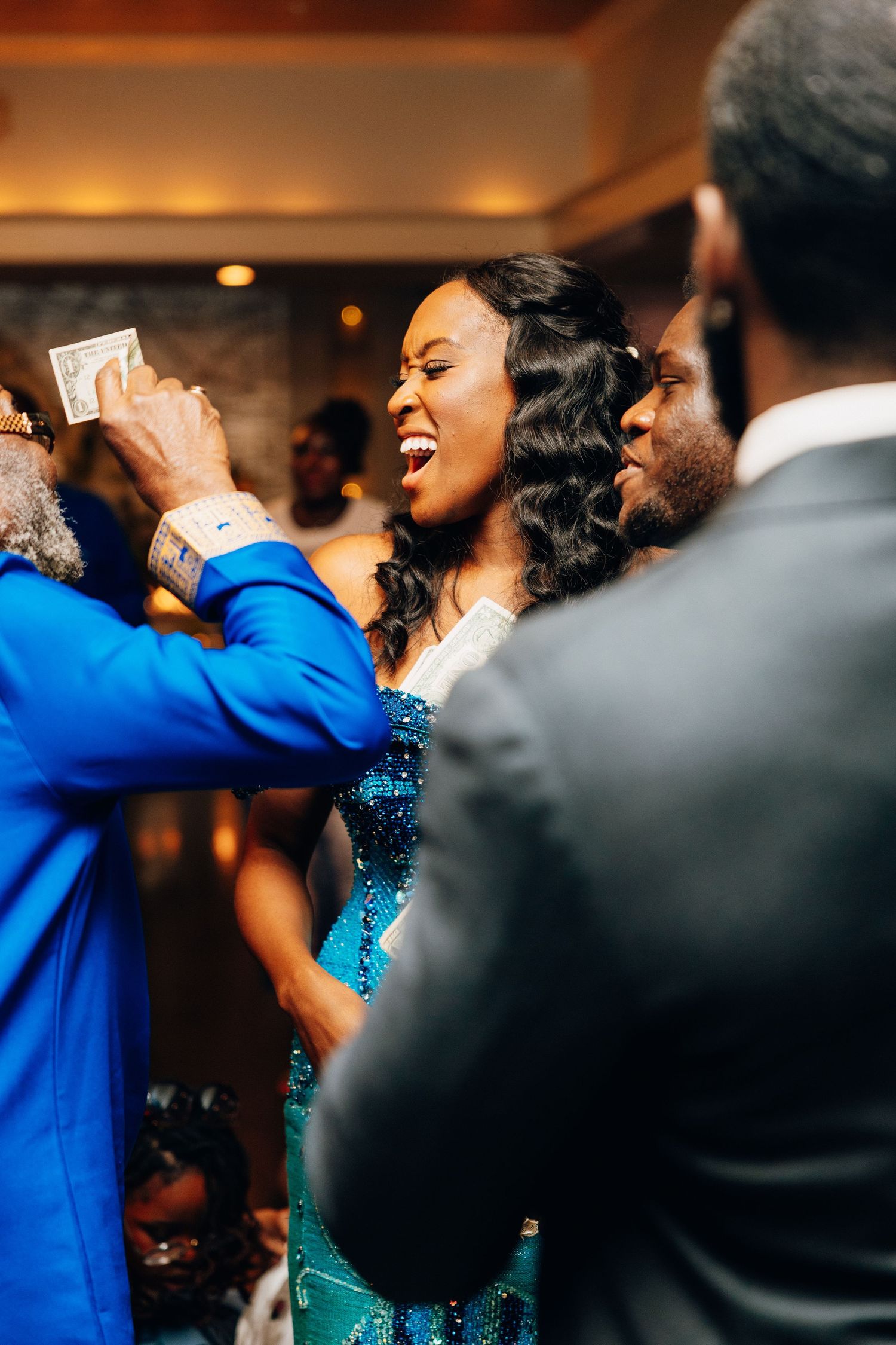 Bride in teal sequin gown laughing joyfully during money dance at wedding.