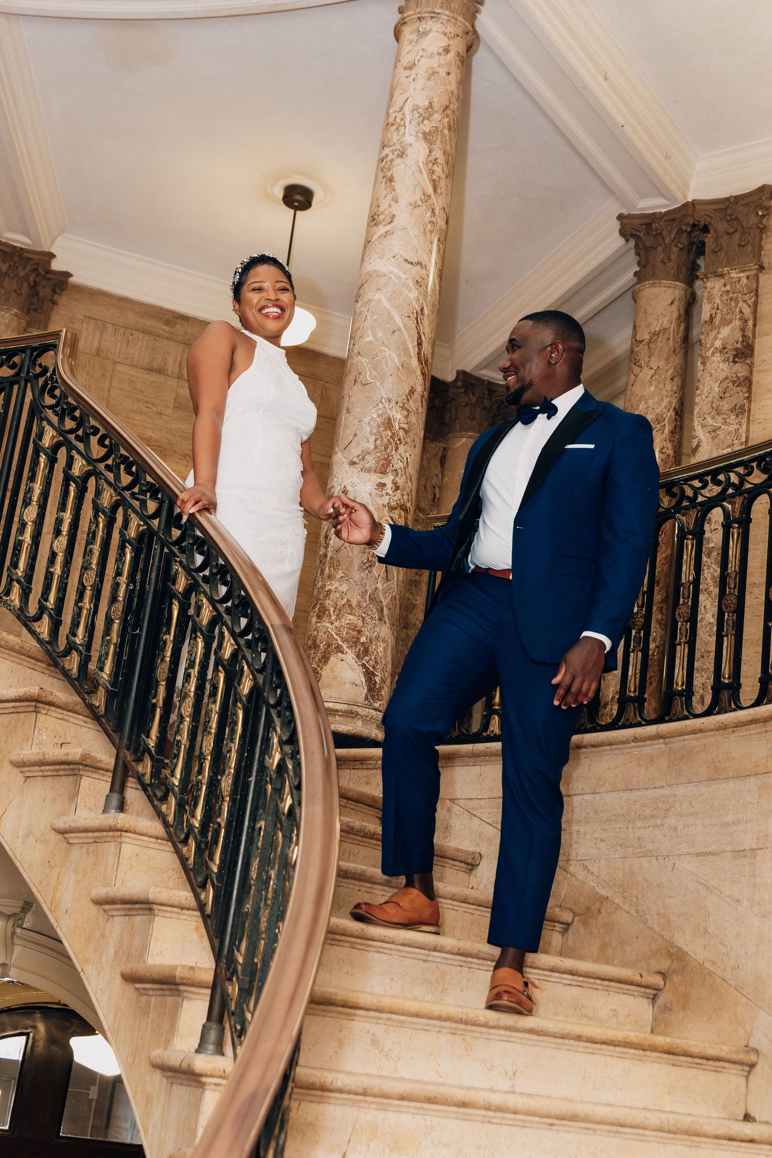 Bride and groom holding hands on elegant marble staircase, she in white halter gown, he in navy tuxedo.