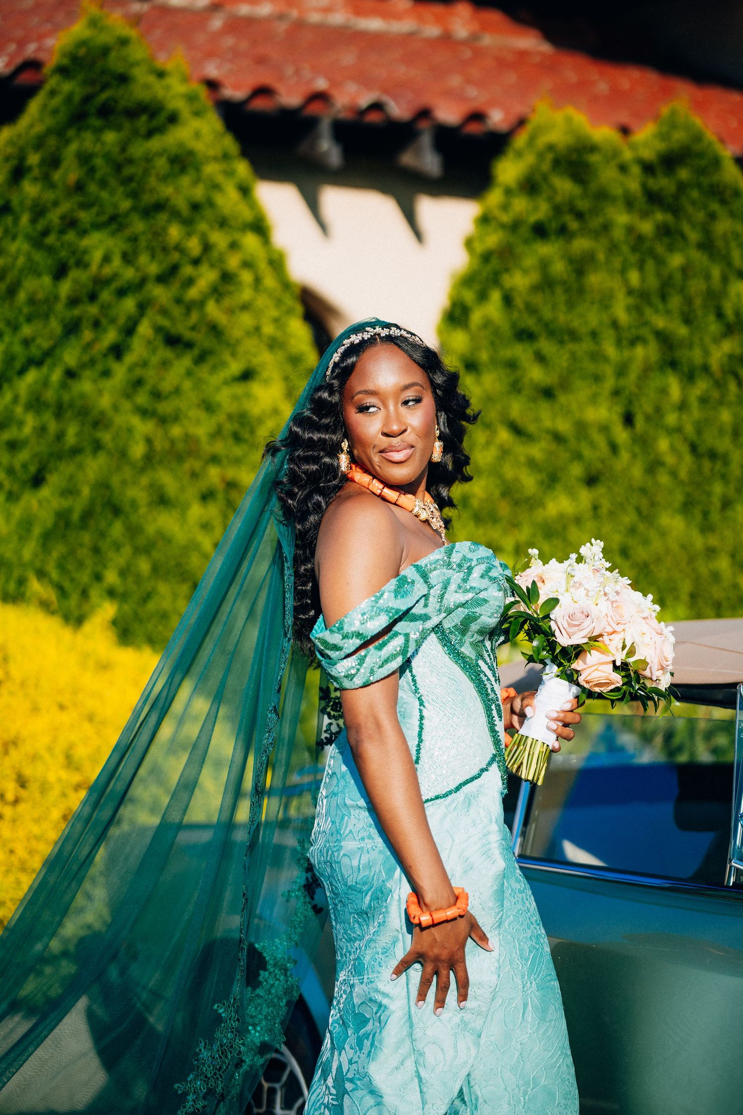 African bride in teal gown with veil poses by vintage car outdoors.