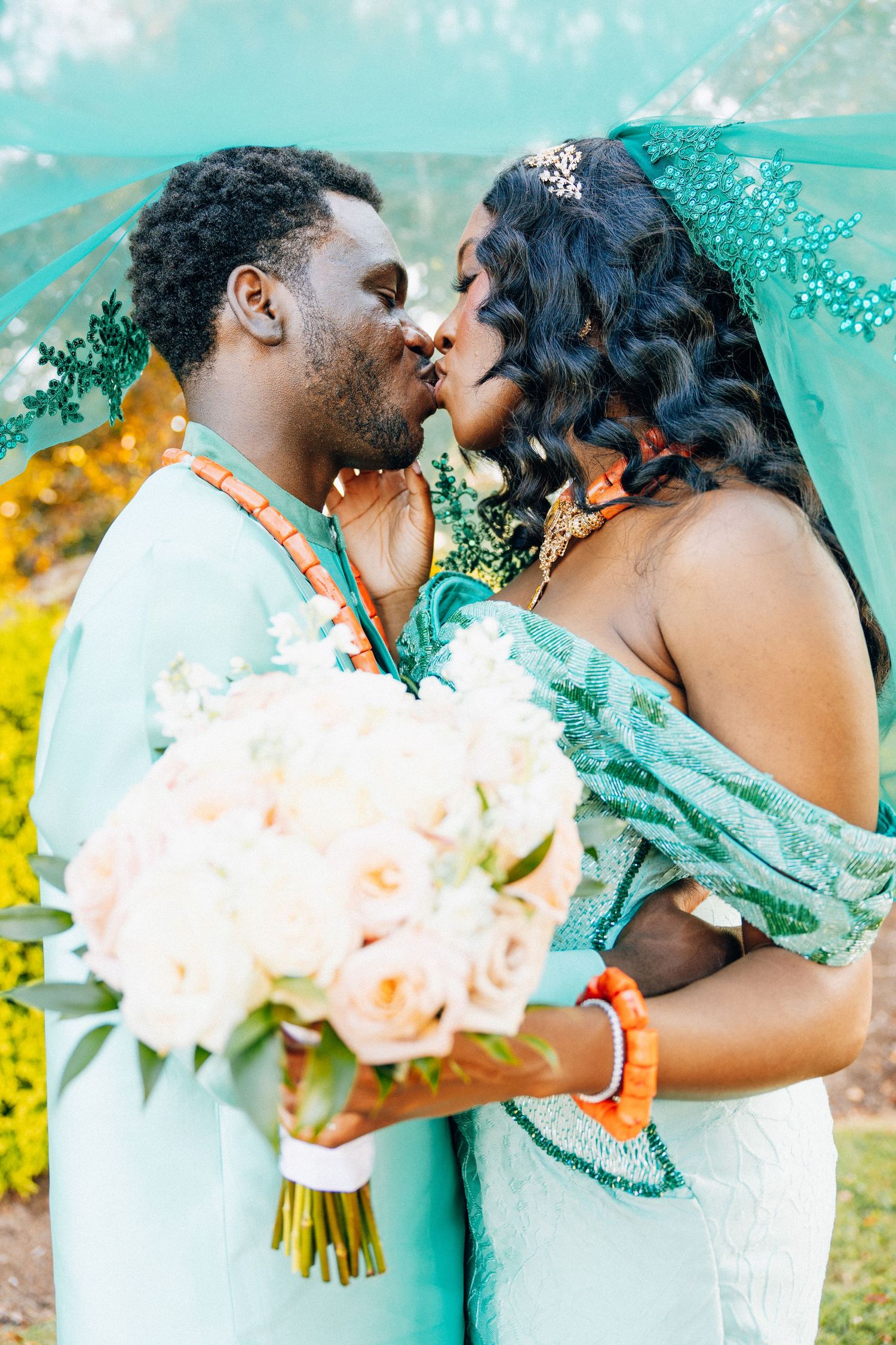African couple sharing a kiss at their traditional Nigerian wedding, wearing matching teal outfits with coral beaded jewelry.