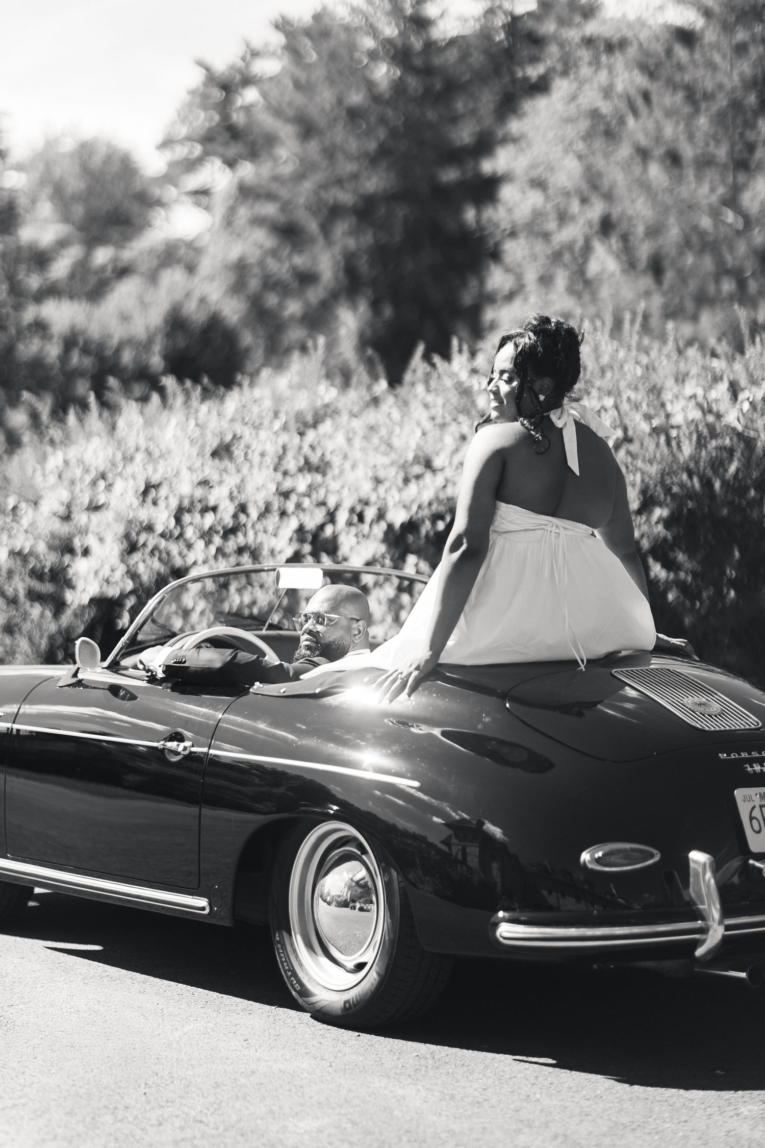 Black and white photo of bride sitting on vintage Porsche 356 hood while groom drives down road.