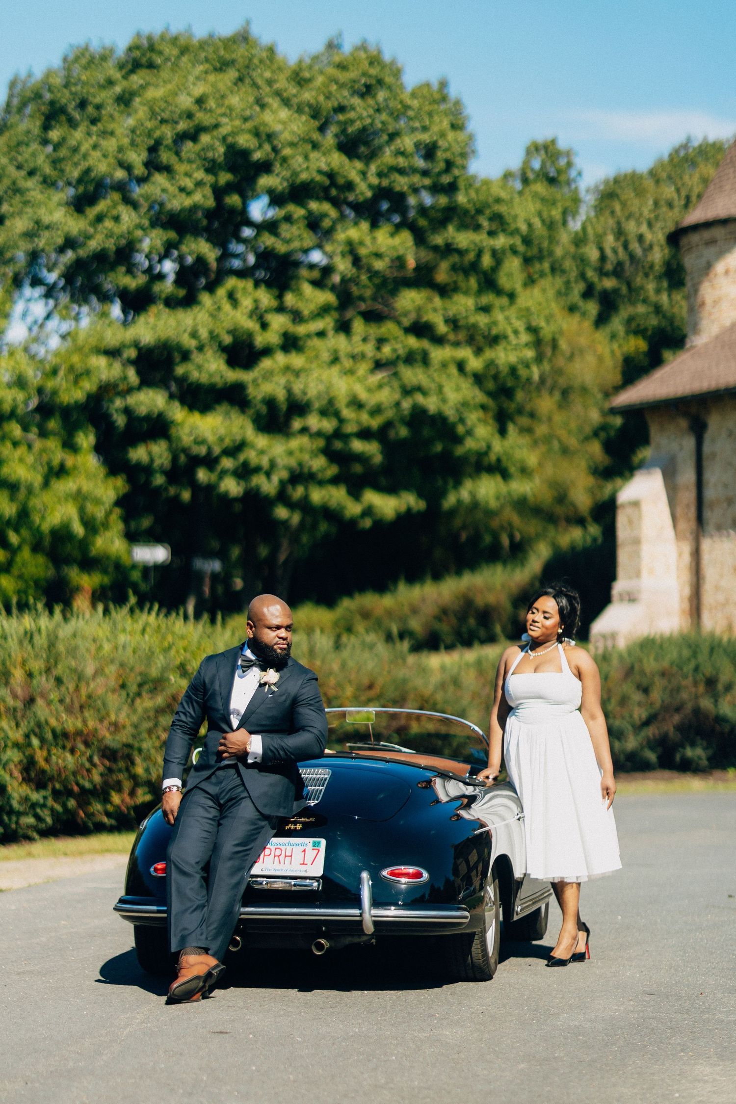 Groom in navy suit leans on vintage black convertible while bride in white dress stands nearby on sunny day.