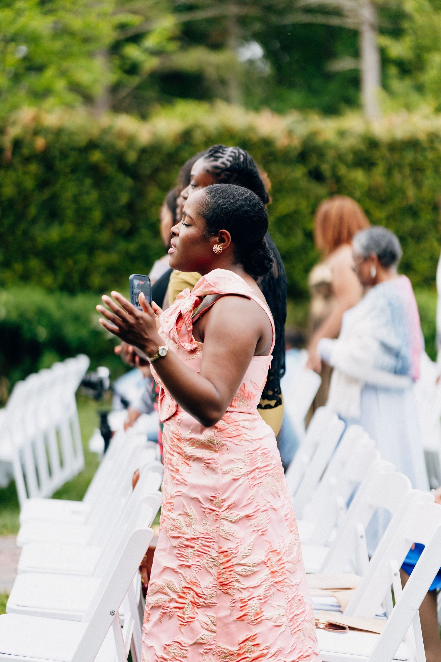 Woman in pink floral dress clapping at an outdoor wedding ceremony with white chairs and lush green trees.