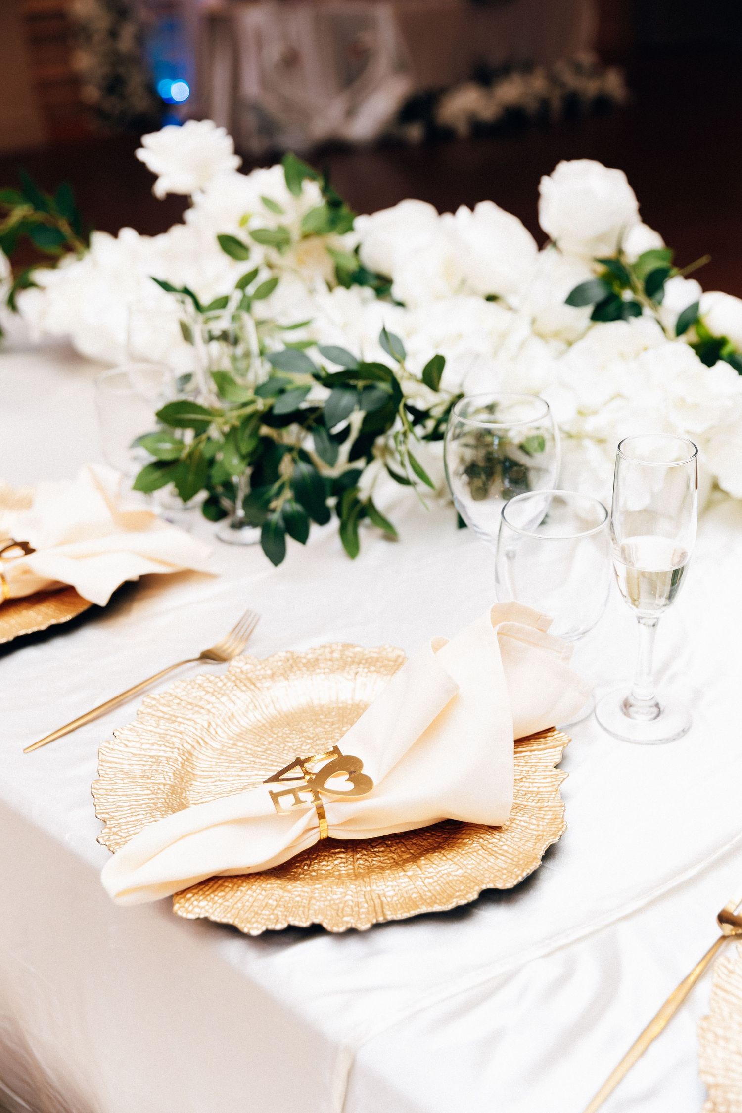 Elegant wedding table setting with gold charger plates, white napkins, champagne flutes, and white floral centerpiece.