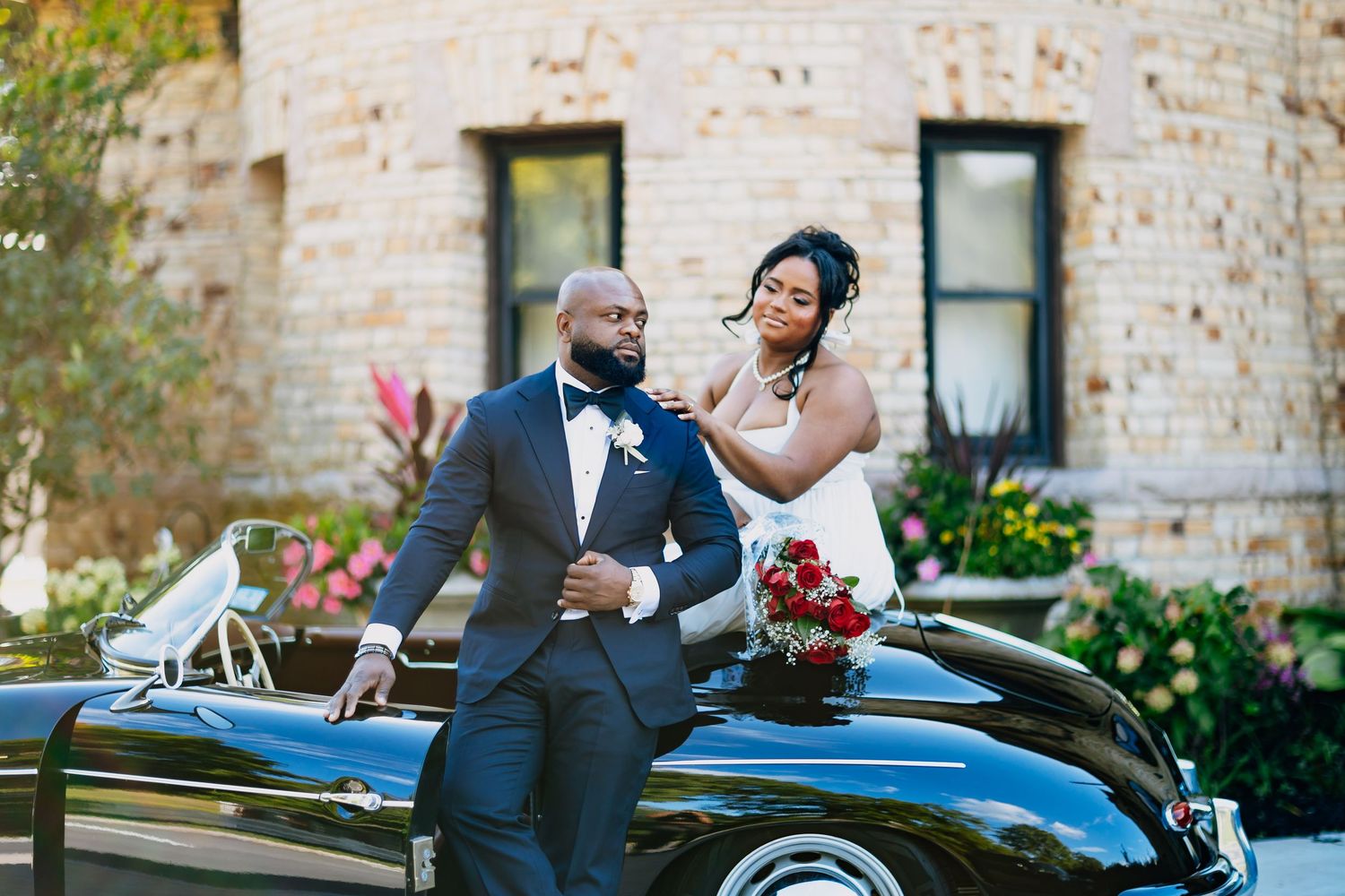 Bride in strapless gown holds red bouquet with groom in navy suit leaning on a classic black convertible car.