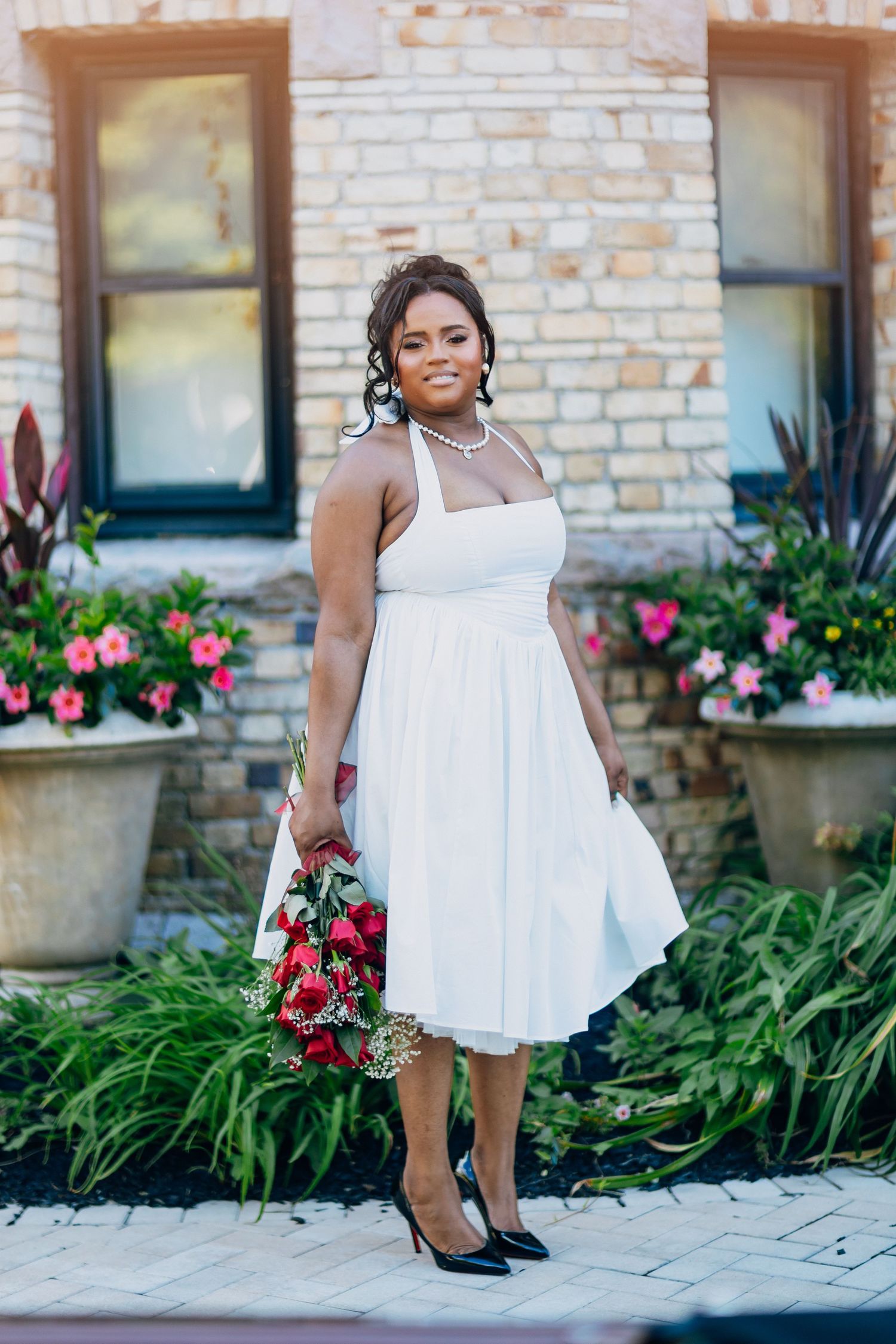 Smiling Black bride in white halter wedding dress holding red rose bouquet outdoors.