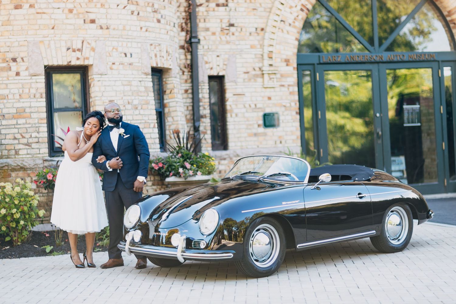 Couple poses beside a vintage black Porsche 356 convertible outside a brick building at their wedding.