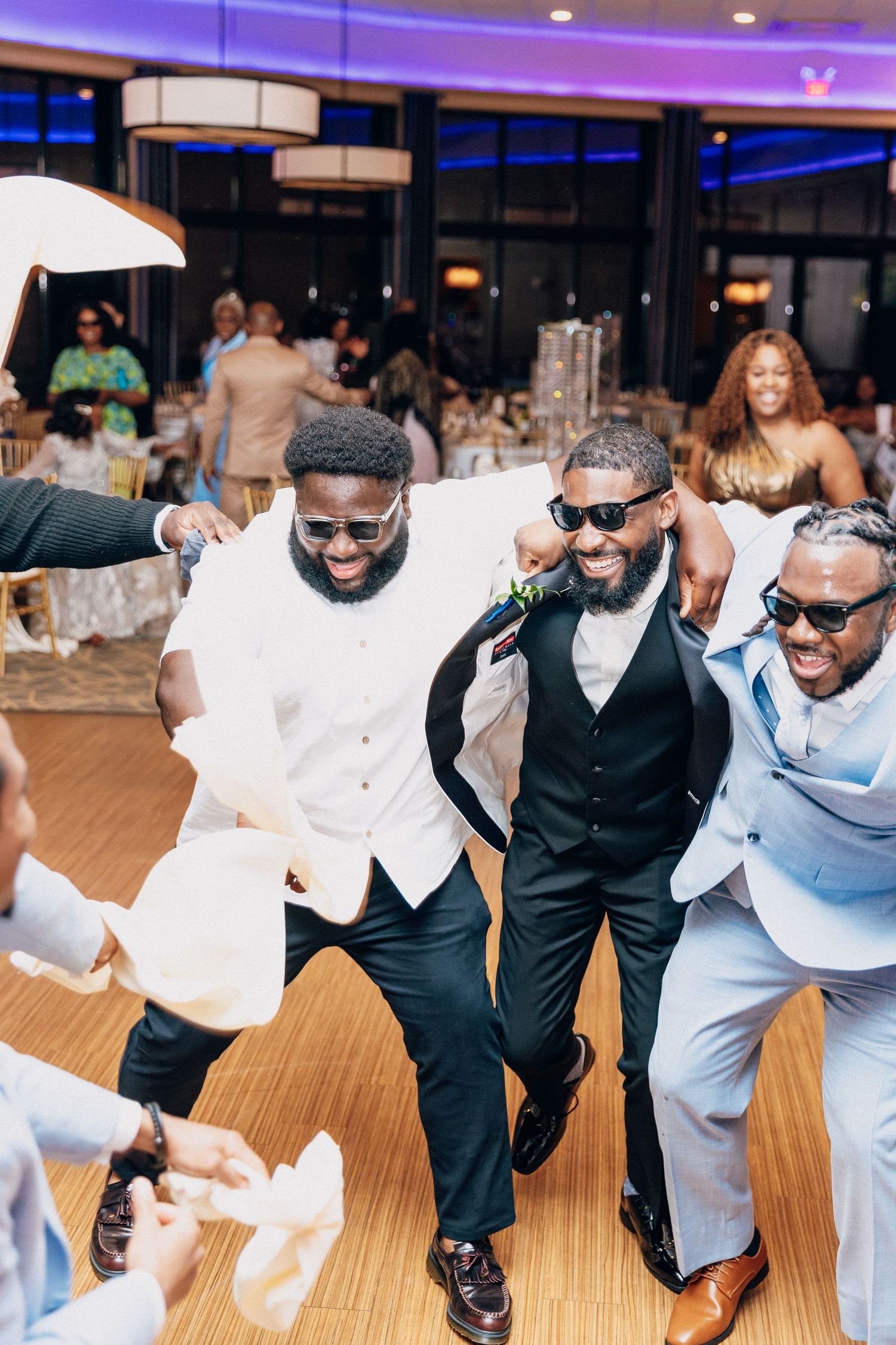 Groom and groomsmen in light blue and black suits dance joyfully at a lively wedding reception.