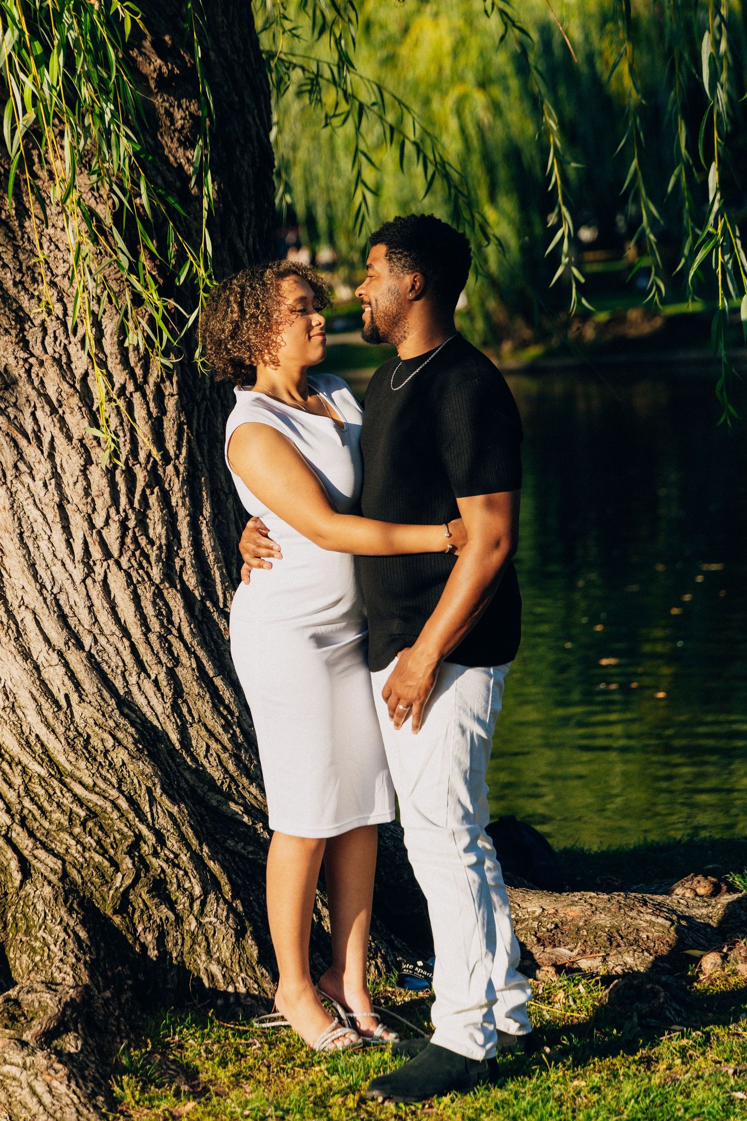 A couple embraces near a willow tree by a pond, woman in white dress and man in black shirt.