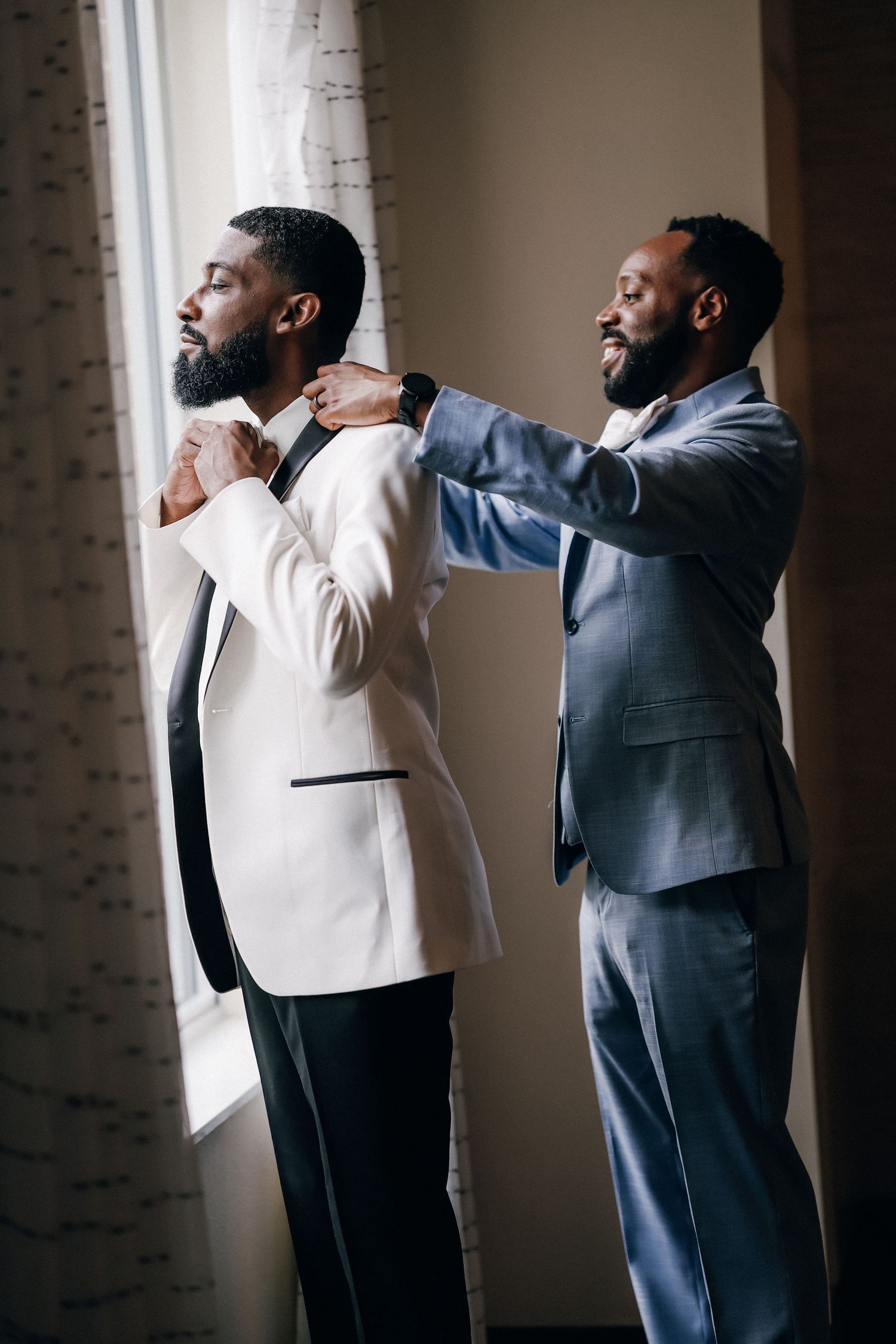 Two Black men in formal suits stand by a window as one helps the other adjust his bow tie.