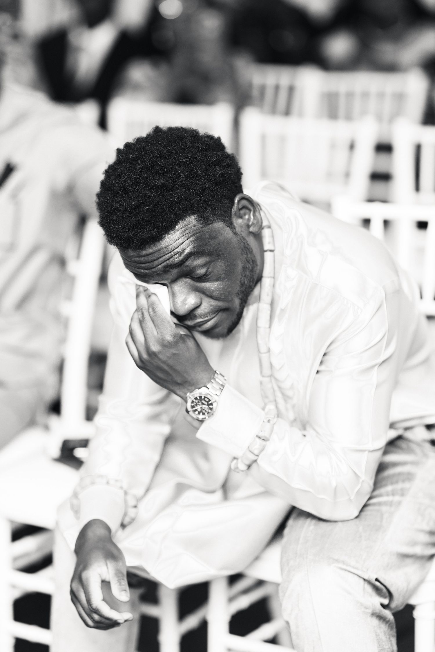 An emotional groom in a white suit wipes tears during a wedding ceremony in black and white photo.