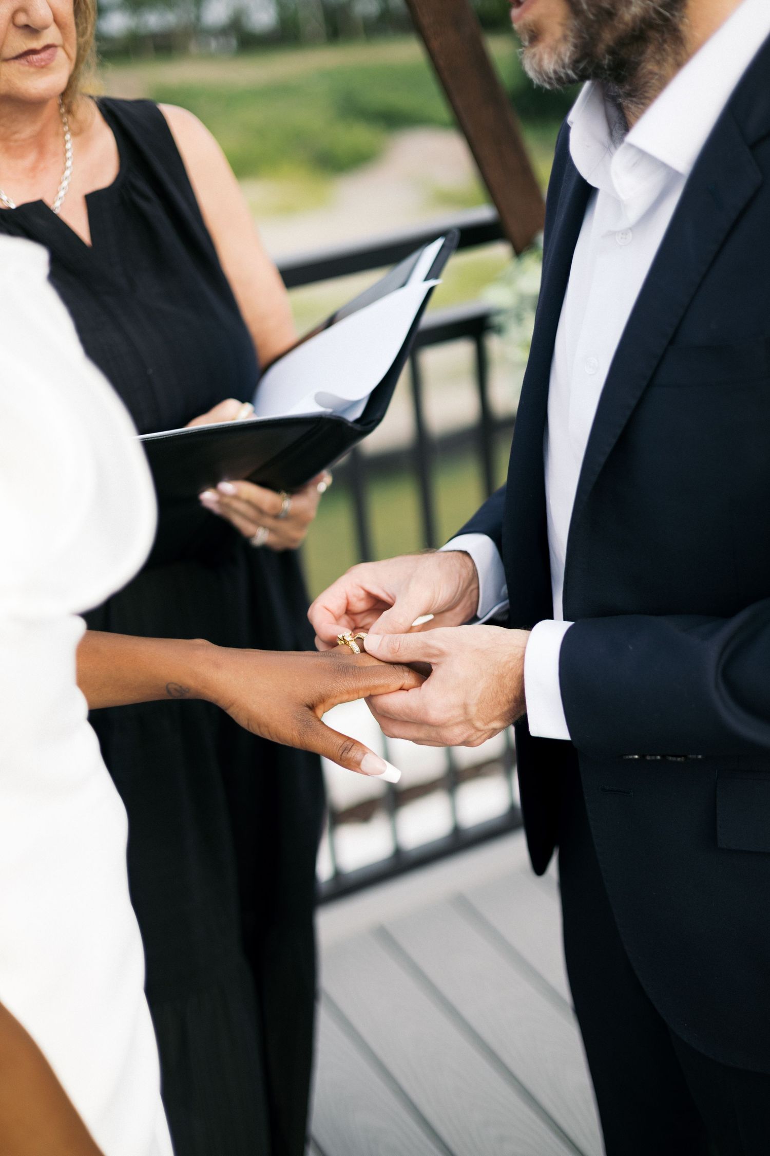 Groom placing a wedding ring on bride's hand during outdoor ceremony with officiant holding book.