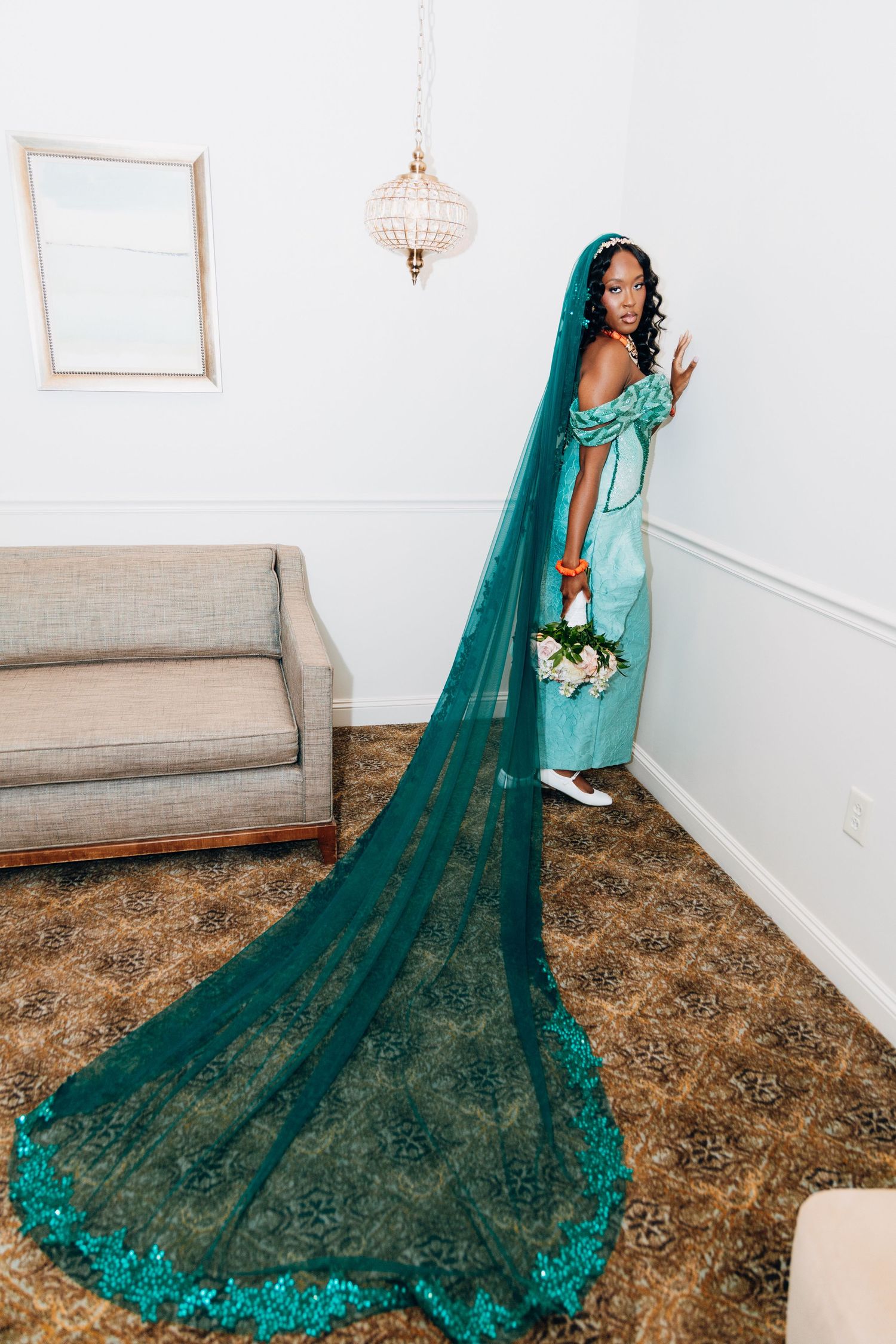 African bride poses in a teal off-shoulder gown with long lace veil and bouquet in elegant room.