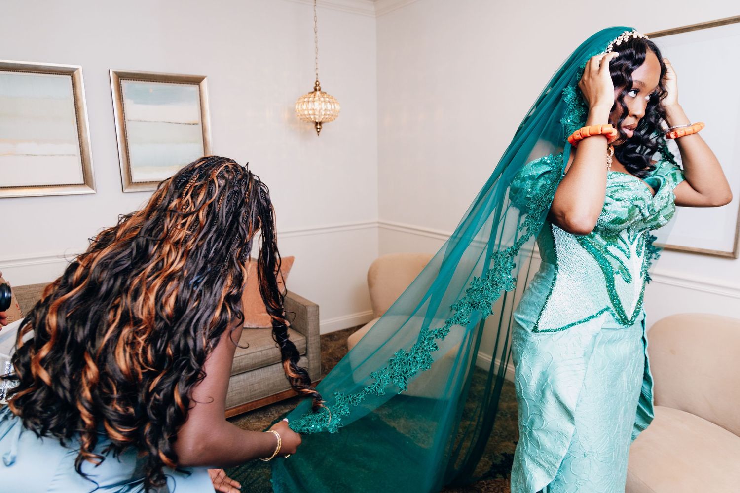 Bride in teal gown adjusts veil while bridesmaid arranges the train in an elegant wedding preparation room.