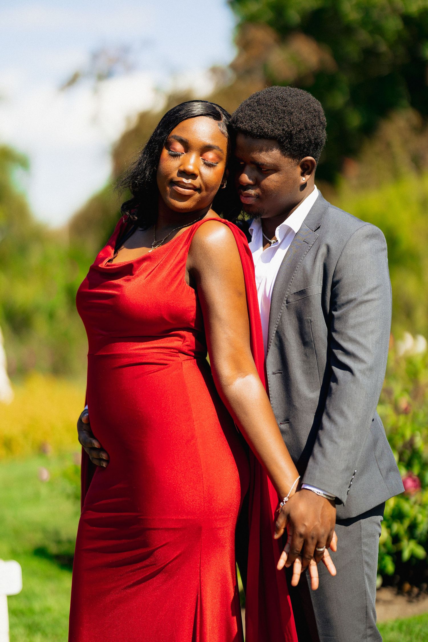 A couple poses in a garden, woman in red gown and man in gray suit holding hands.