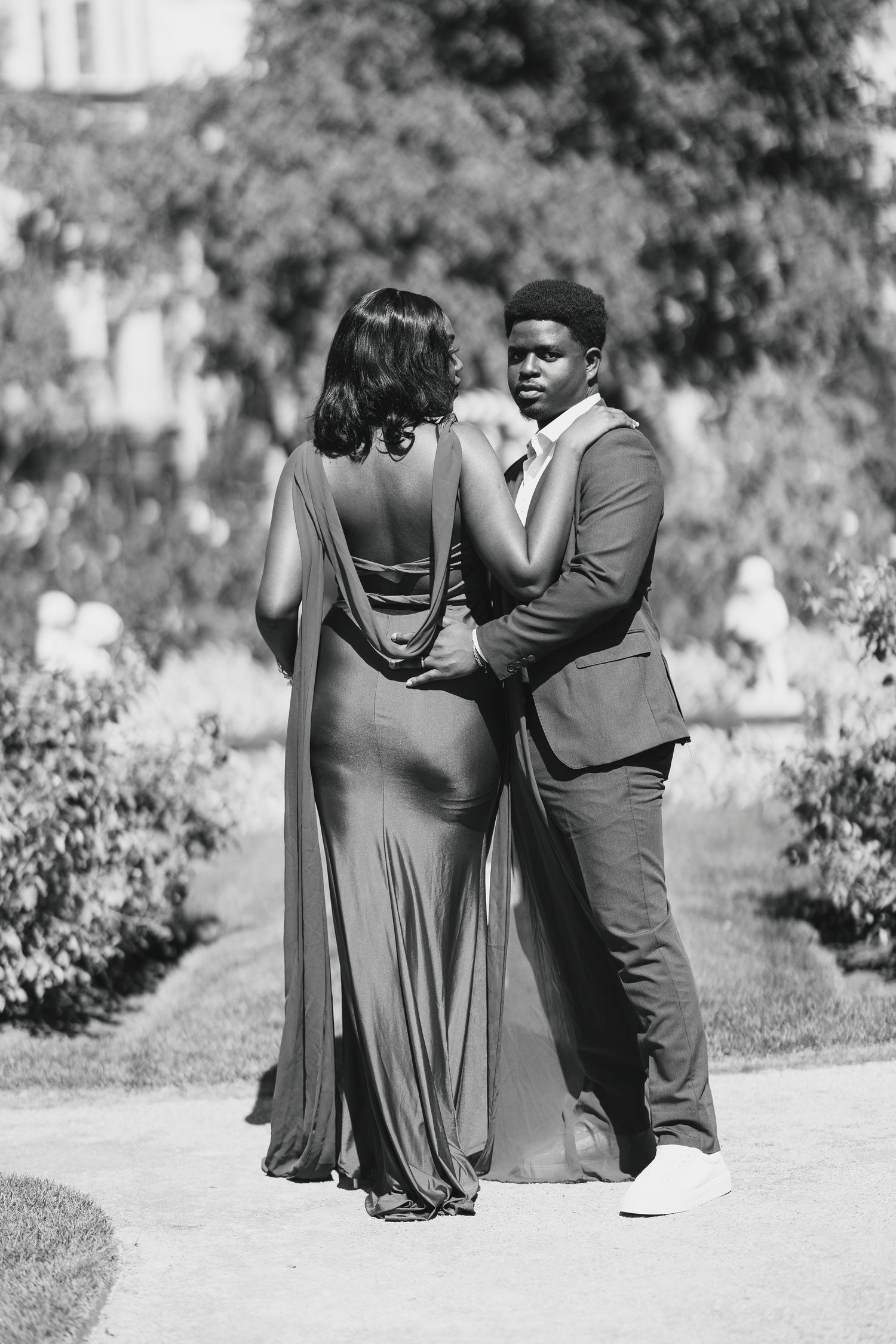 Black and white photo of an elegant couple in formal attire posing in a lush garden setting.