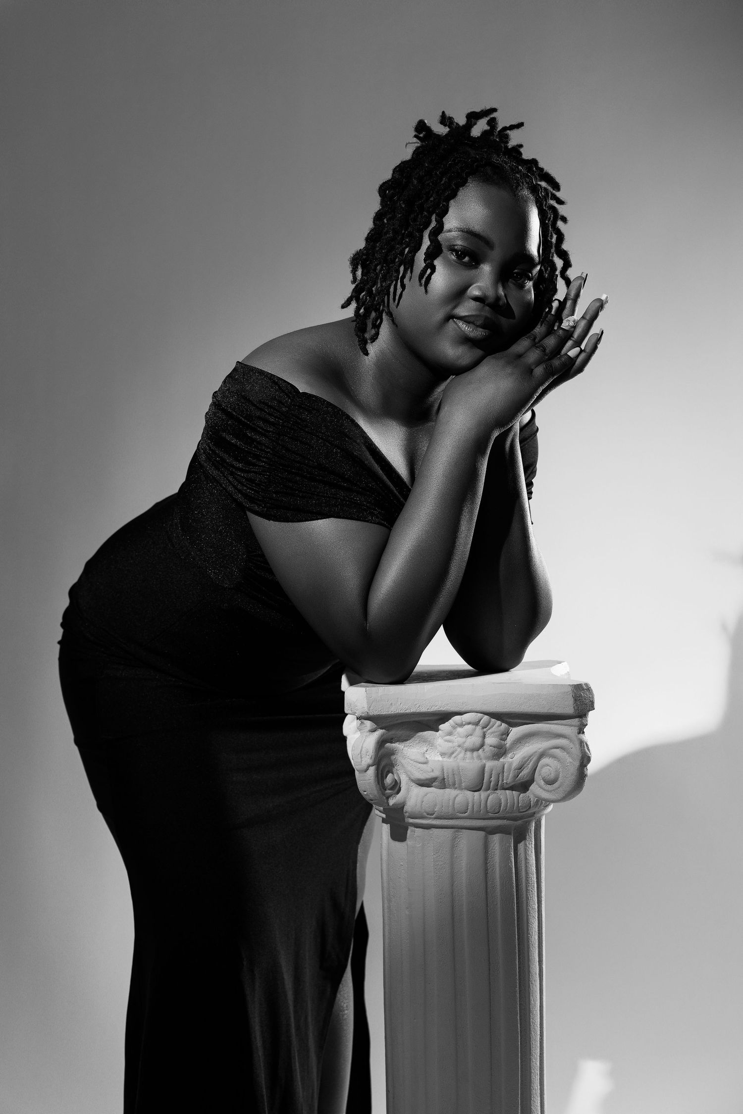 Black and white portrait of a woman in an off-shoulder dress leaning gracefully on a decorative white column.