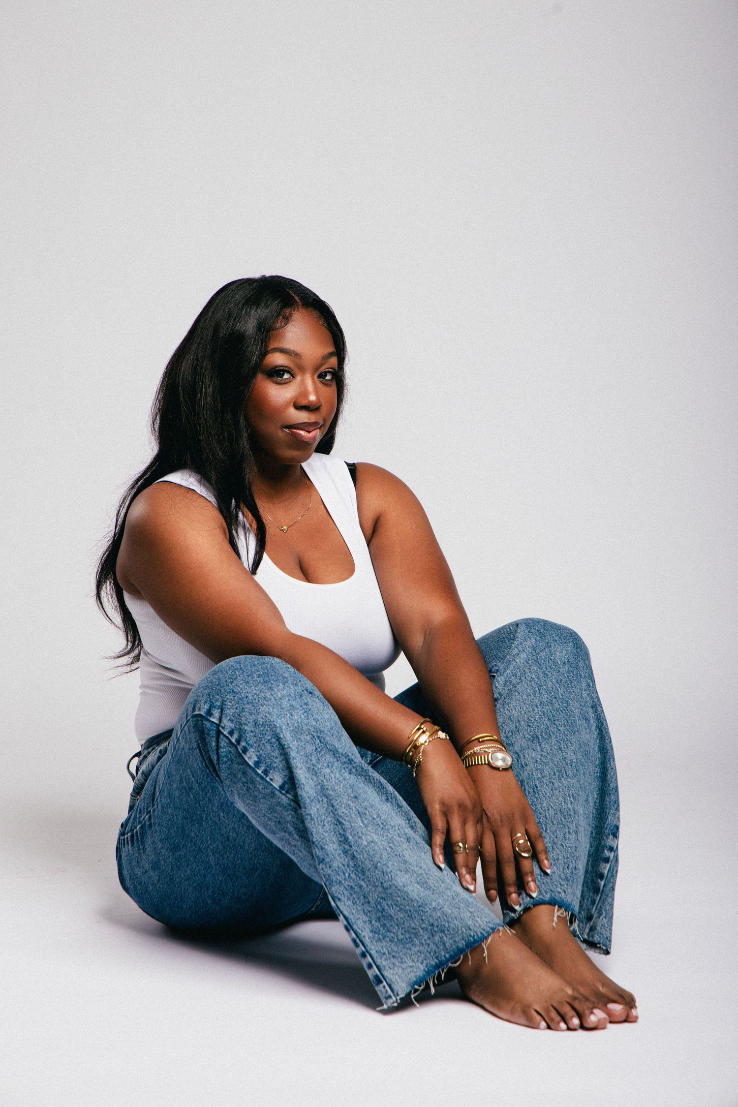 Young Black woman sitting on floor in white tank top and wide-leg jeans, wearing gold jewelry, barefoot, studio shot.