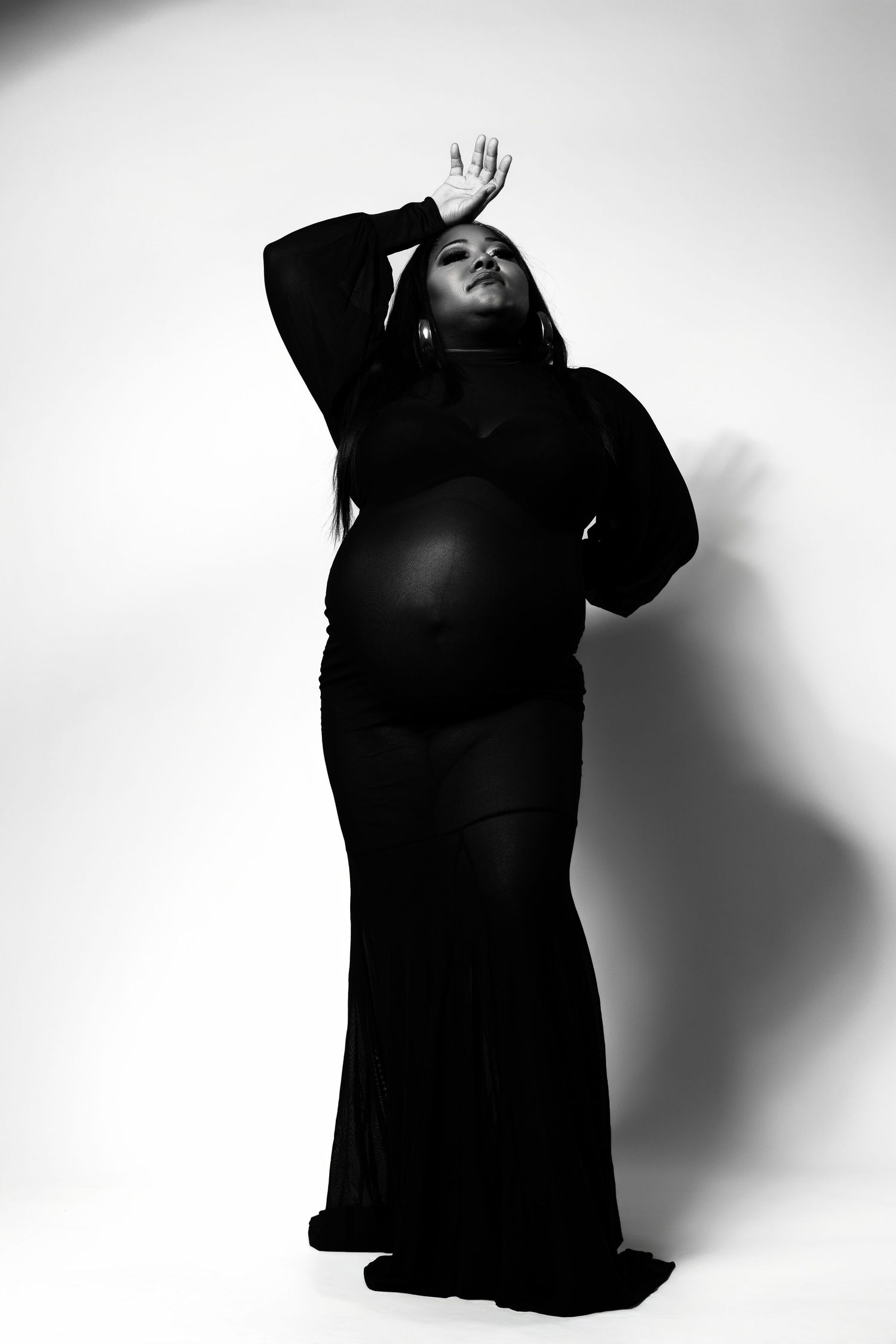 Black and white photo of a pregnant woman in a flowing black gown posing gracefully with one hand raised above her head.