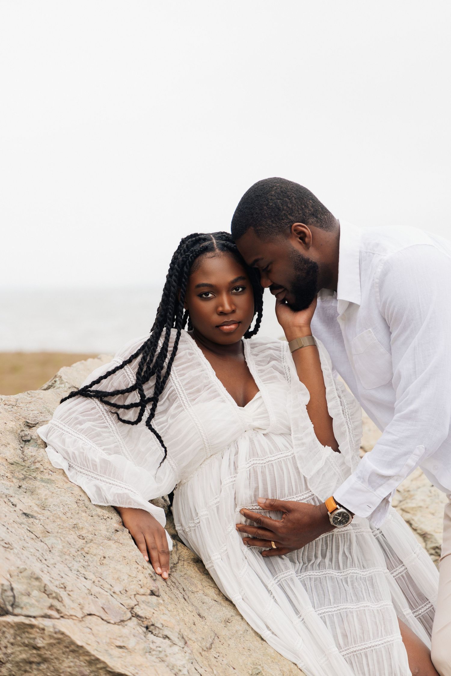 Black couple in matching white outfits sharing intimate moment on rocks, pregnant woman cradling baby bump during maternity shoot.