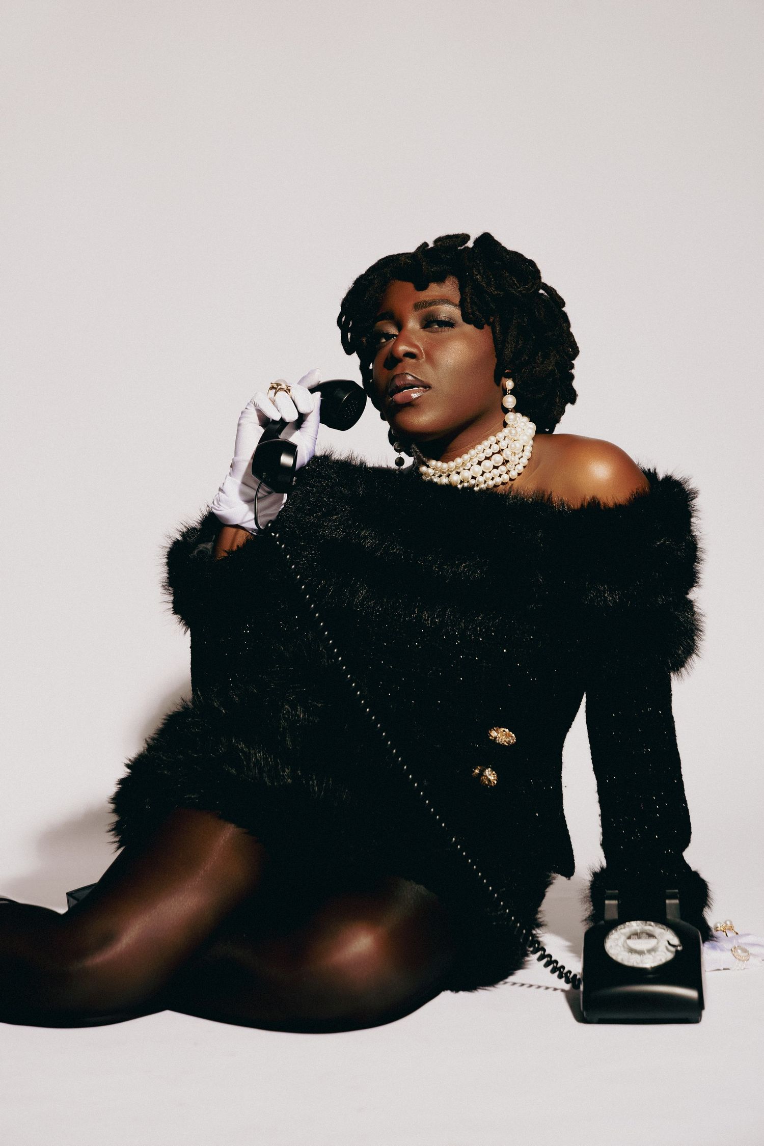 A glamorous Black woman in a black fur off-shoulder outfit holds a vintage rotary phone, wearing pearl jewelry and white gloves.