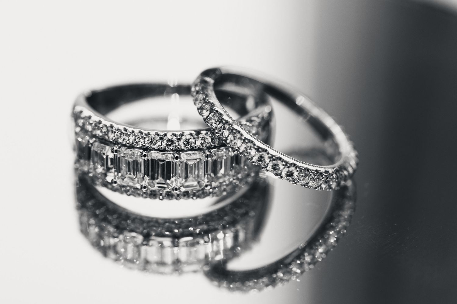 Two elegant diamond wedding rings with baguette and pavé settings reflected on a glossy surface in black and white.