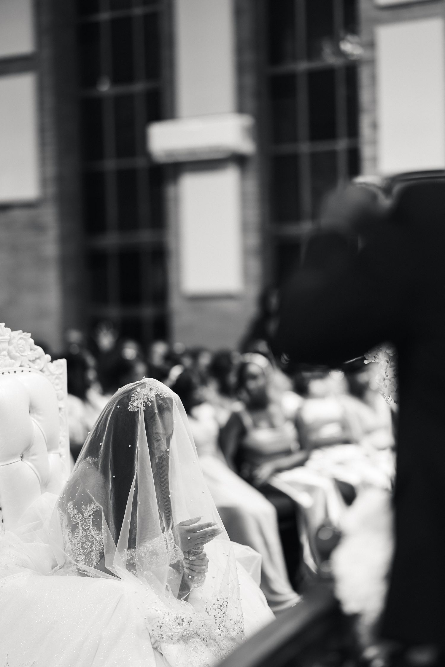 Black and white photo of a bride in lace veil seated during wedding ceremony with guests in background.