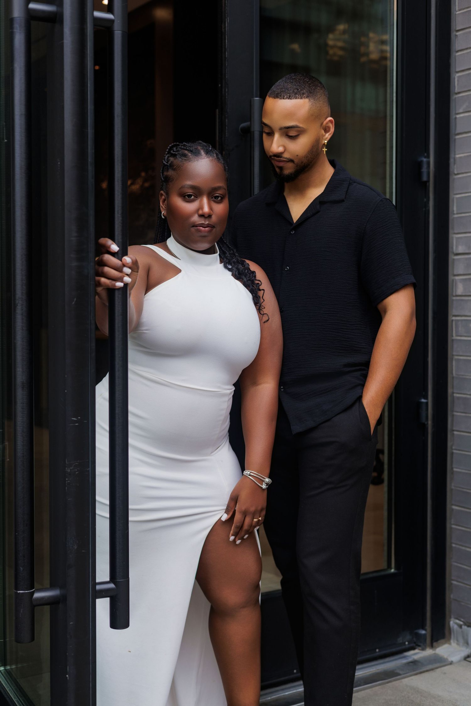 A stylish couple poses in a doorway, woman in white dress and man in black outfit, against a modern backdrop.