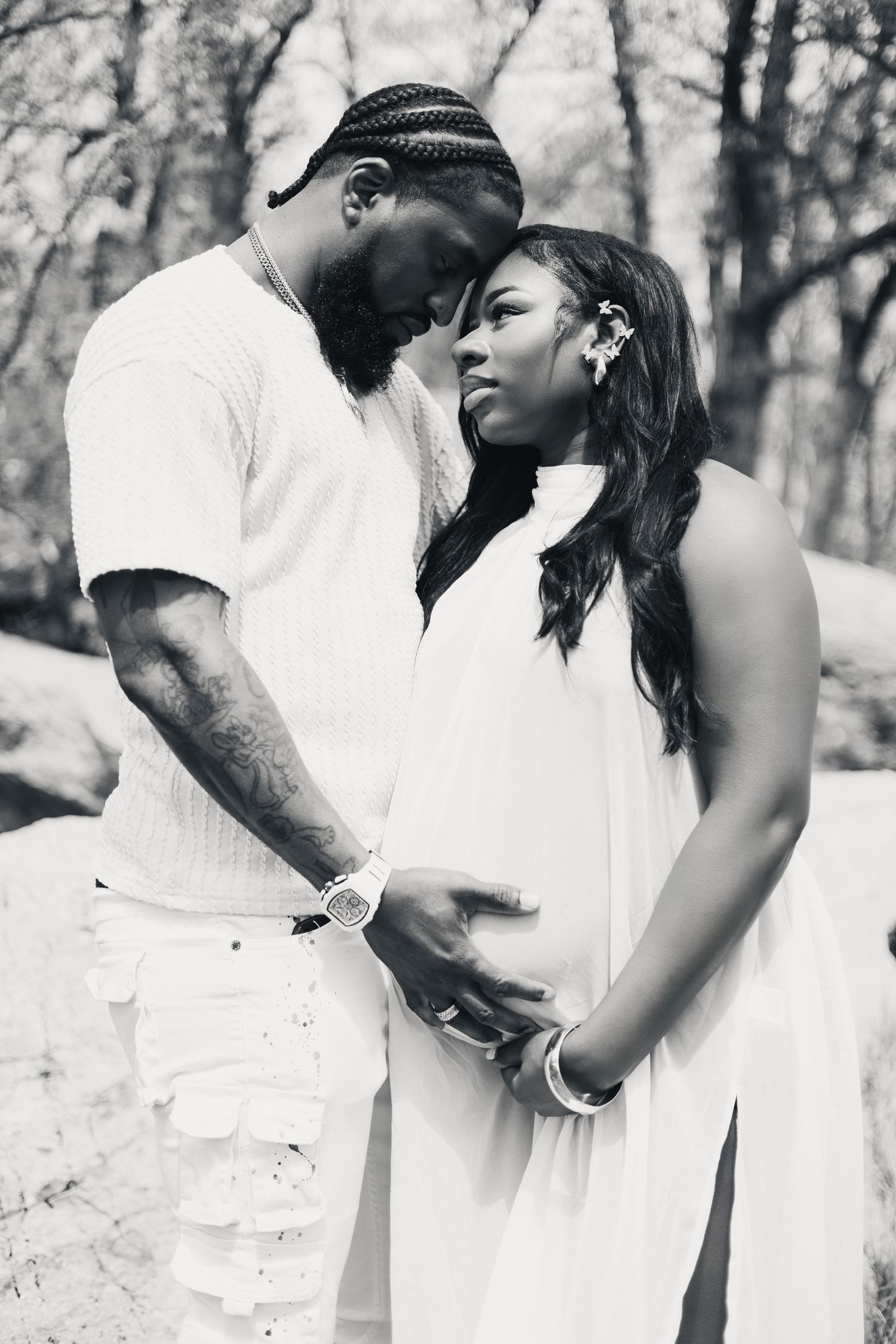 Black and white maternity photo of a couple in white outfits tenderly touching baby bump outdoors.