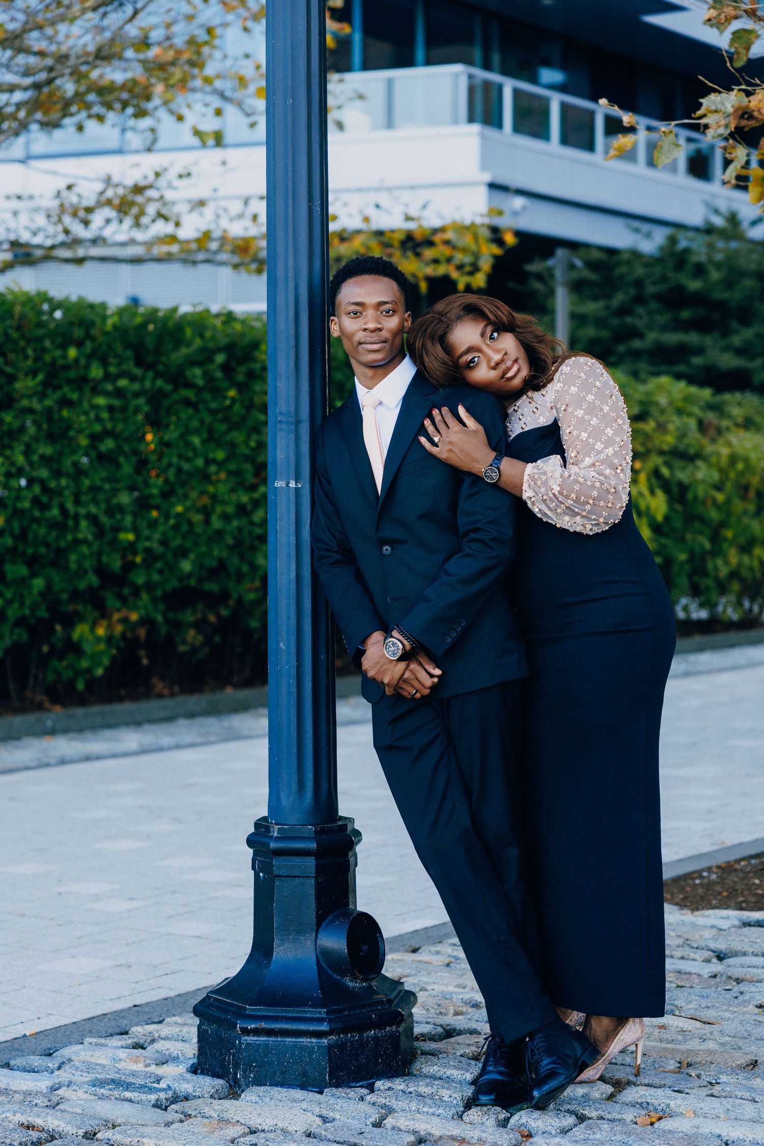 Elegant couple posing by a blue lamppost outdoors, man in dark suit, woman in beaded top leaning on his shoulder.