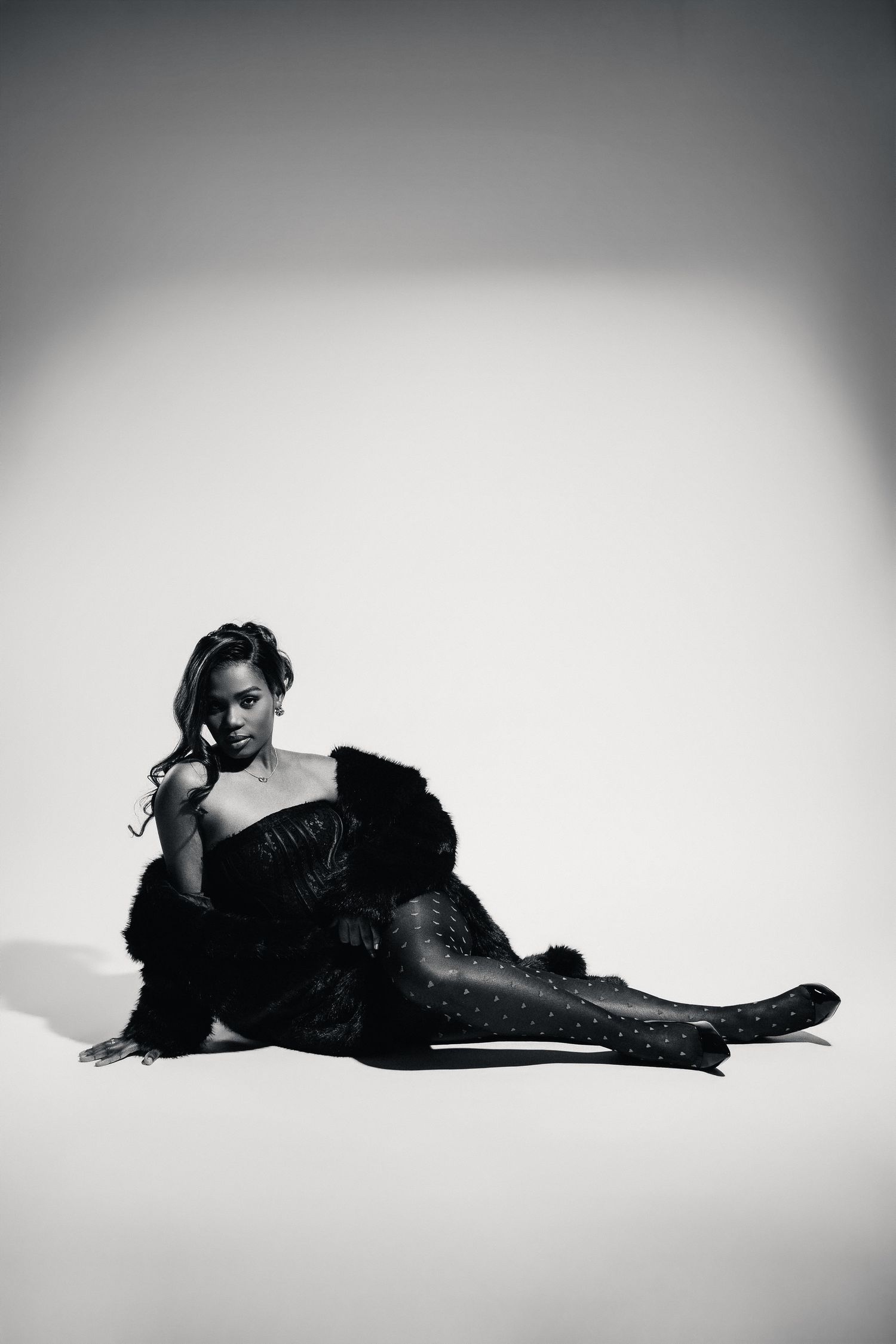 Black and white photo of a woman in a fur coat and studded tights posing on the floor.