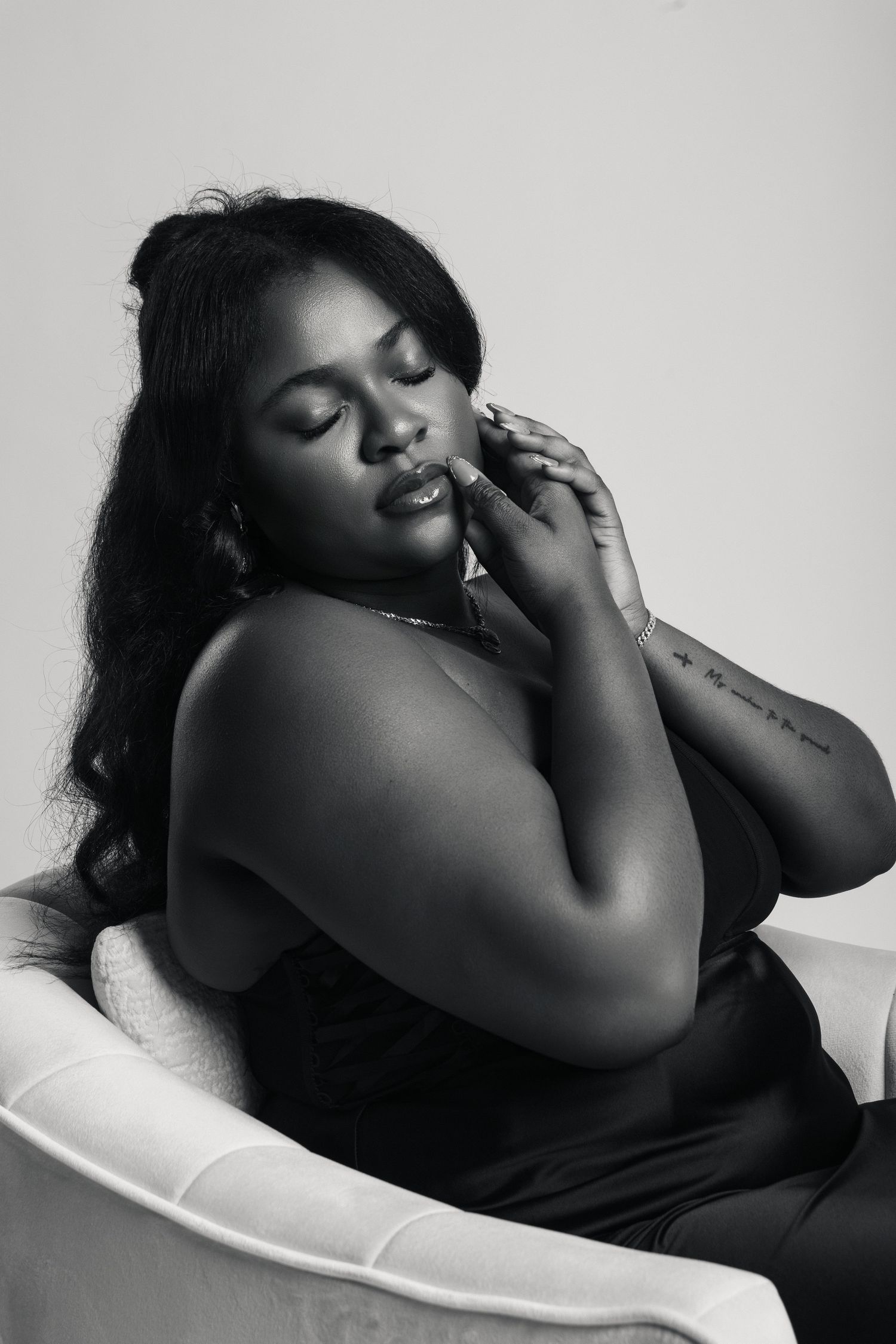 Black and white portrait of a woman sitting in a chair with eyes closed, hand near face, wearing a black top.